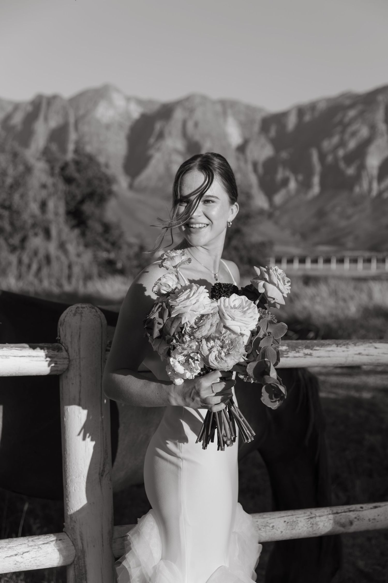 Smiling bride in white gown holds bouquet by rustic fence with mountain backdrop in black and white photo.