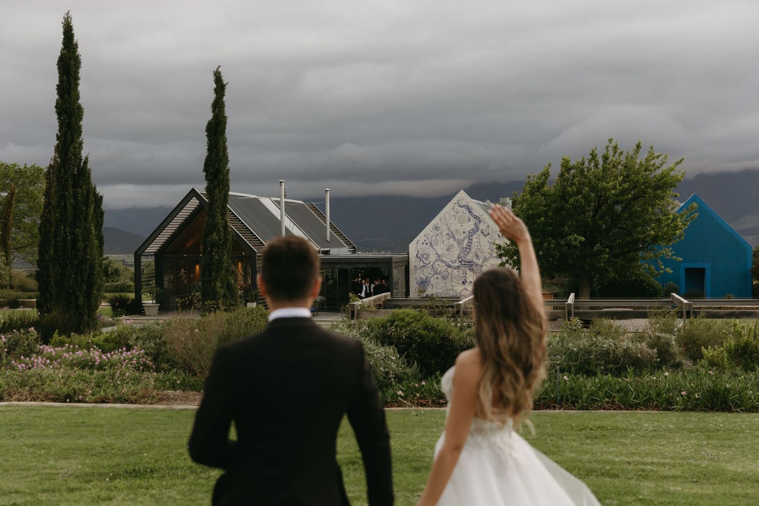Bride and groom walk through lush garden toward rustic venue with cypress trees and mountains under dramatic cloudy sky.