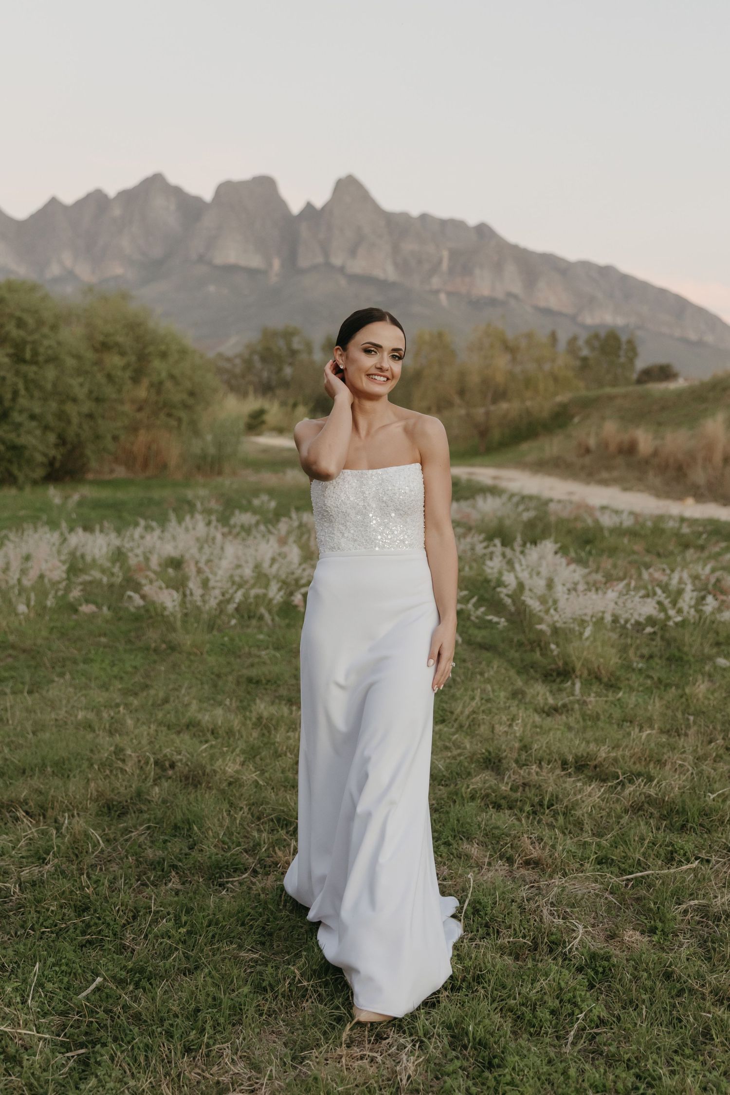 Bride in white strapless gown standing in a green field with dramatic mountain backdrop at sunset.