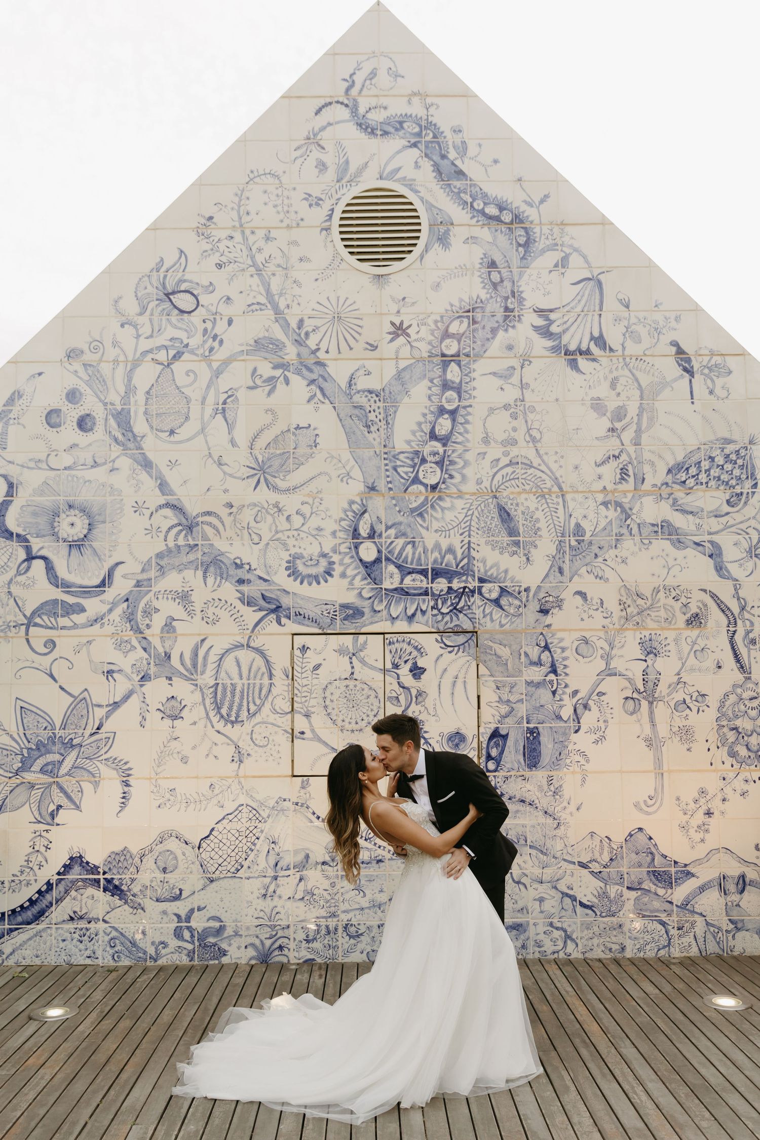 Bride and groom kissing in front of a stunning blue and white floral tile mural on their wedding day.