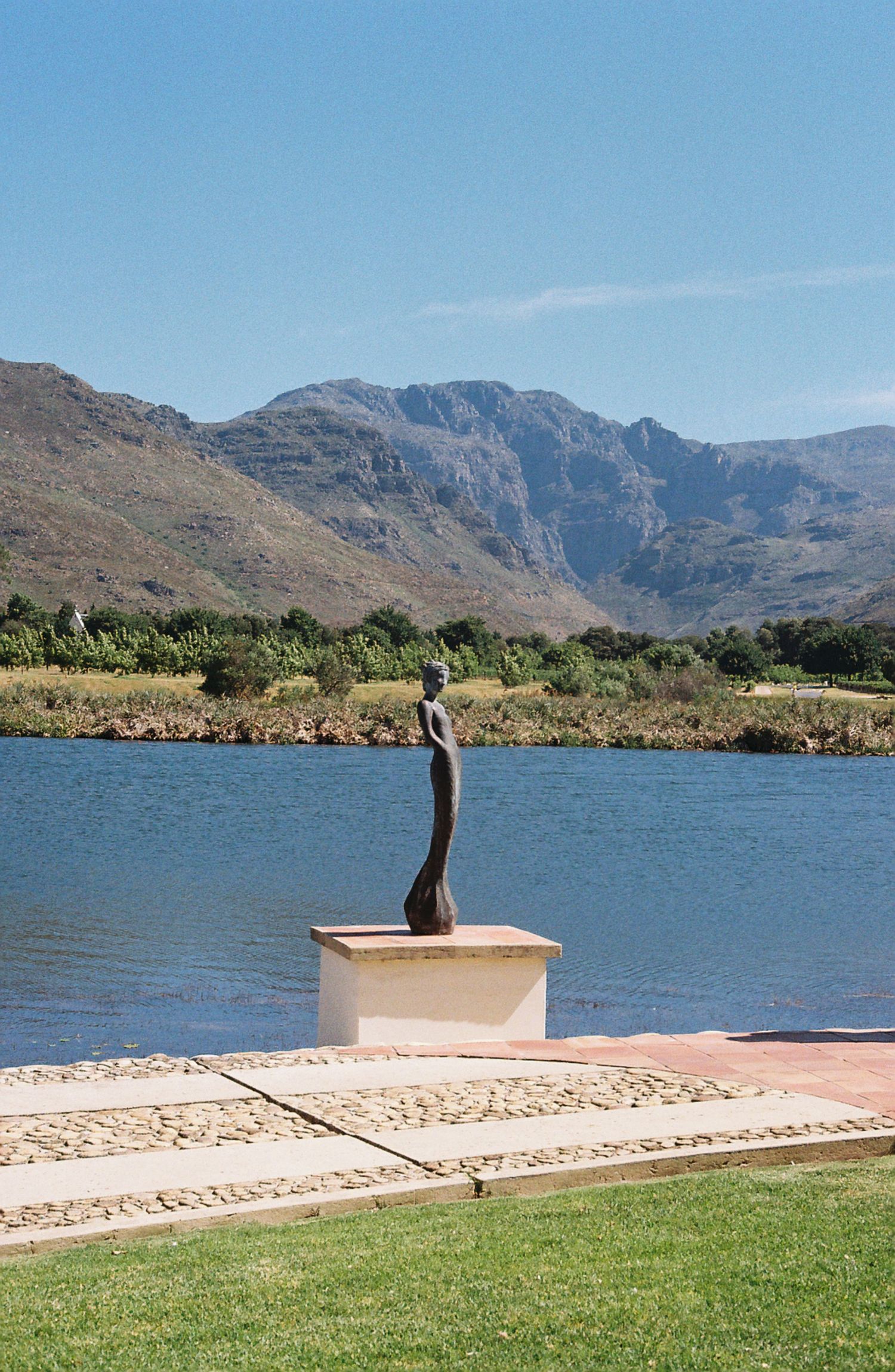 A sleek modern sculpture stands on a pedestal by a lake with dramatic mountain peaks in the background.