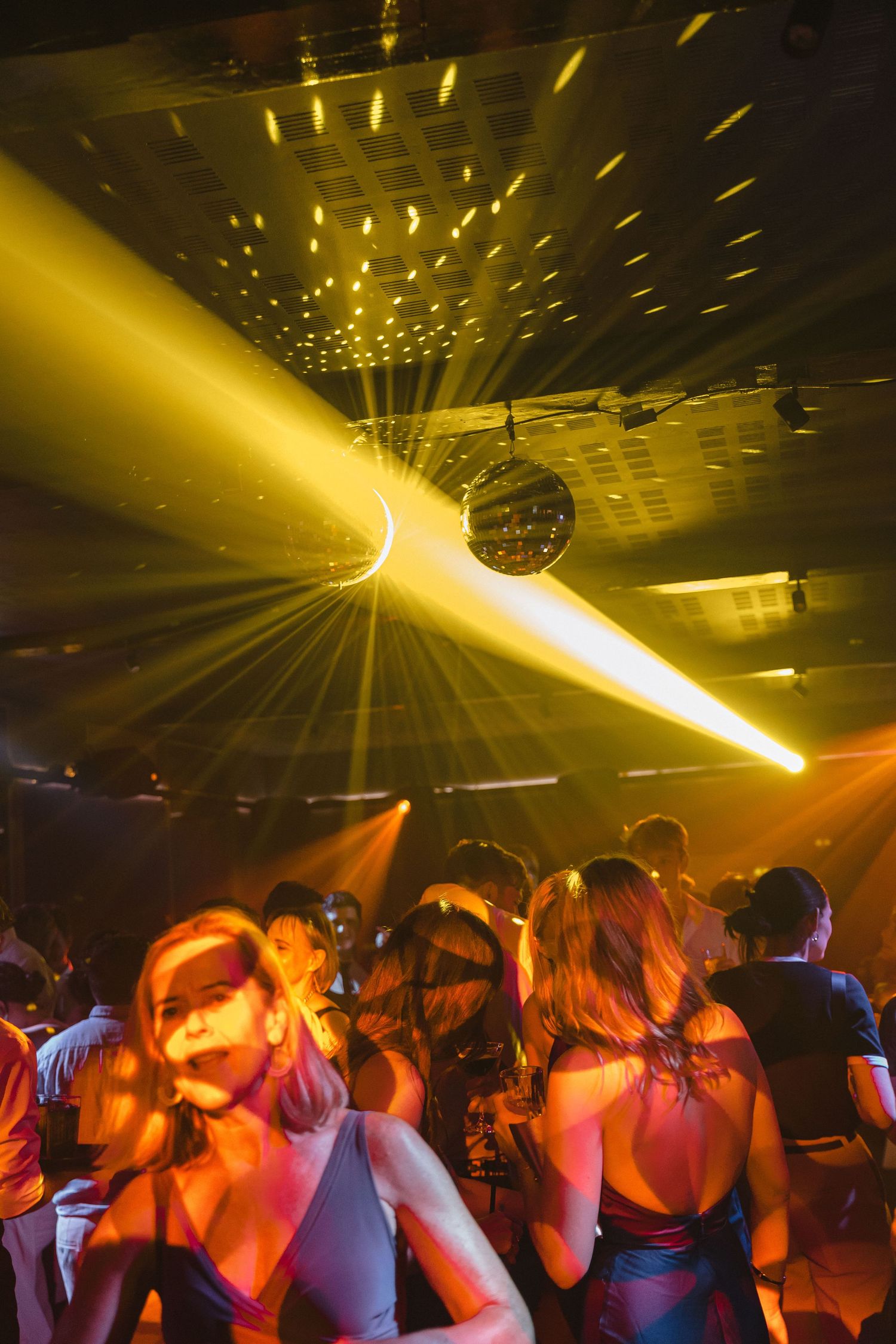 A crowded nightclub dance floor illuminated by yellow spotlights and disco lights while people dance in silhouette.
