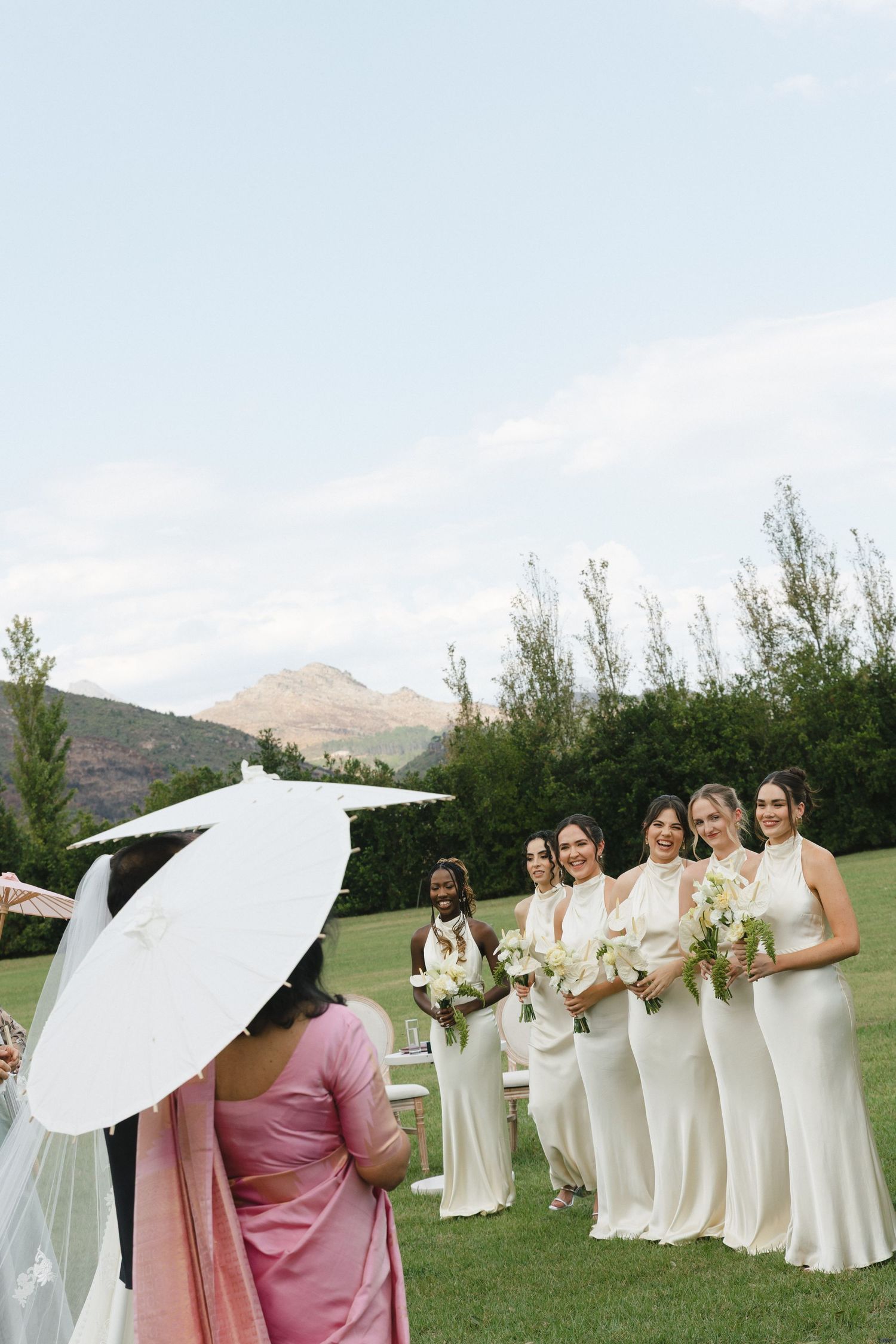 Wedding party members in cream dresses hold white parasols while posing outdoors with mountains in the background.