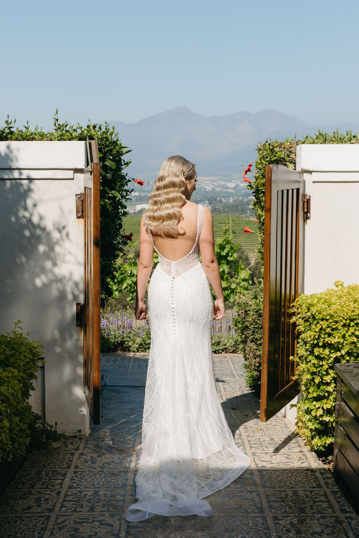 Bride in white lace backless gown with button detail stands between garden gates with mountain views.