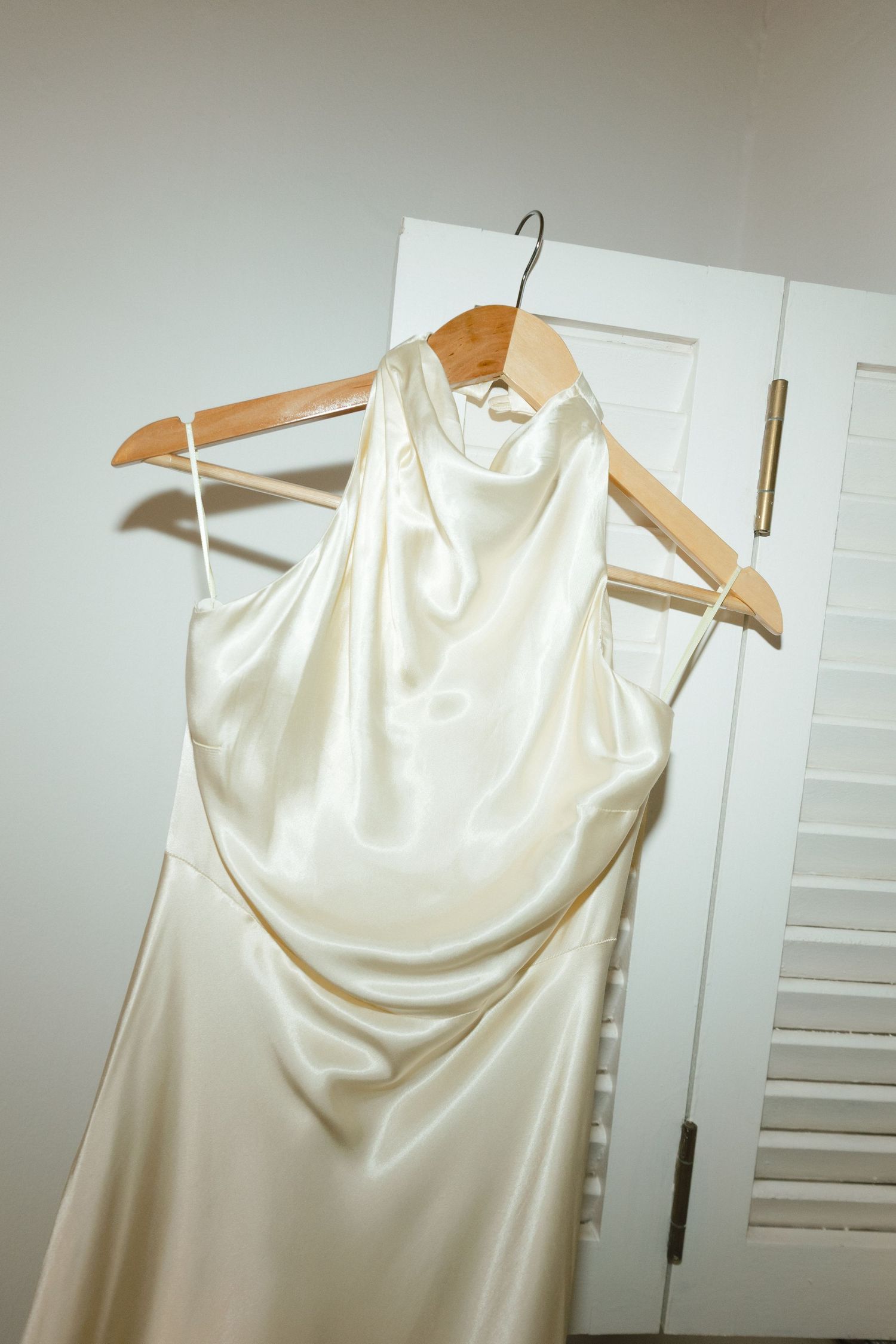 Elegant white satin wedding dress hanging on wooden hangers against light background.