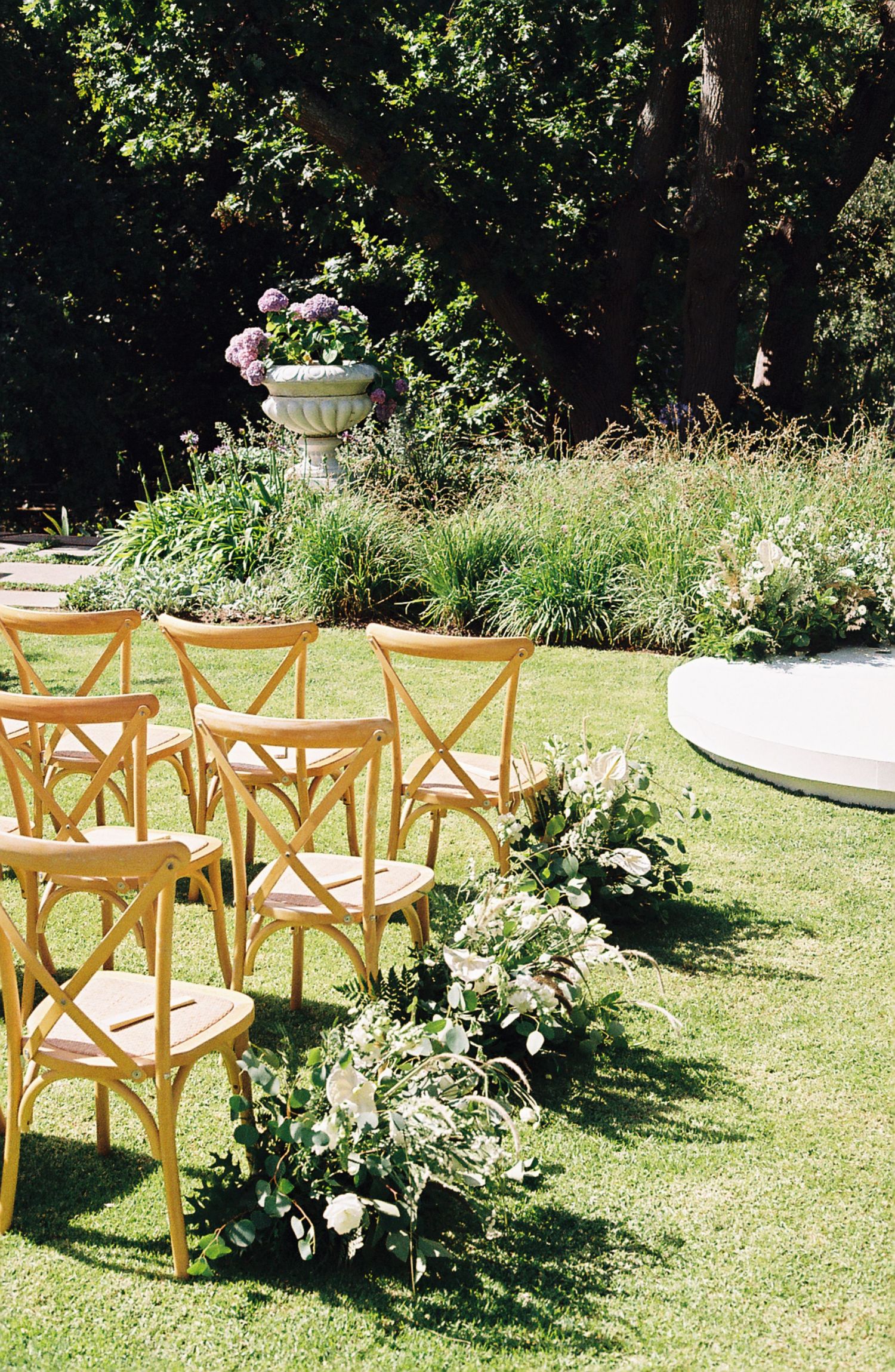 Wooden cross-back chairs arranged on a sunny lawn for an outdoor event with white floral decorations.