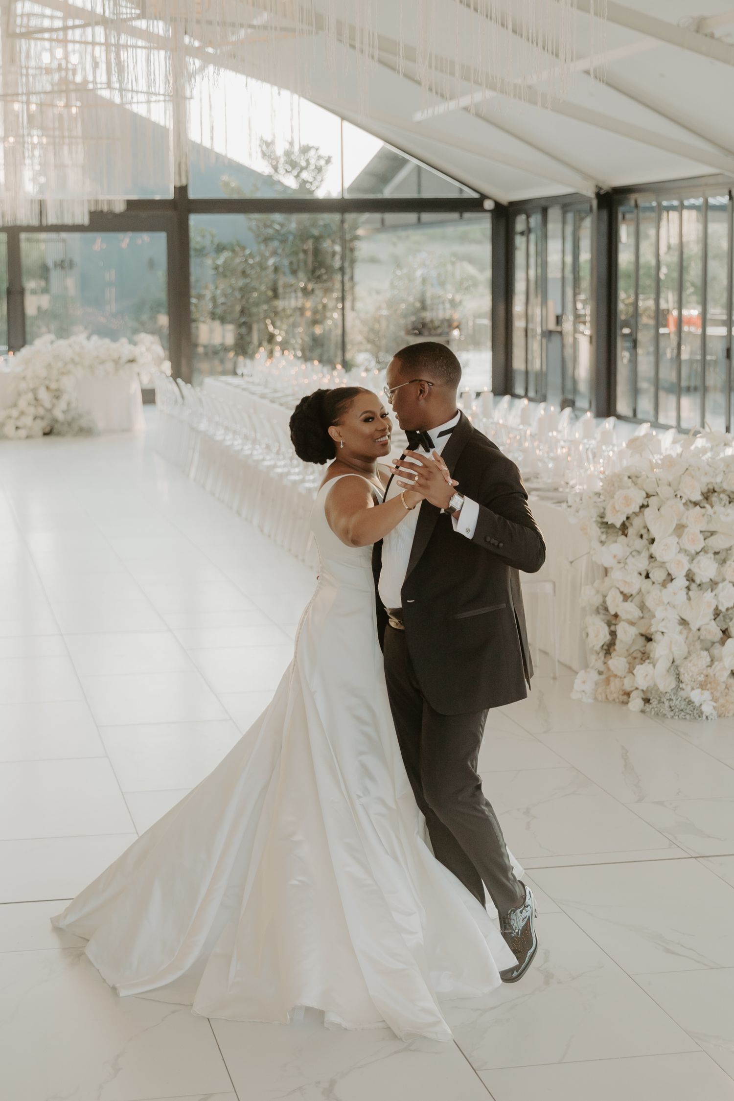 Bride and groom share their first dance in an elegant white wedding venue adorned with floral arrangements.