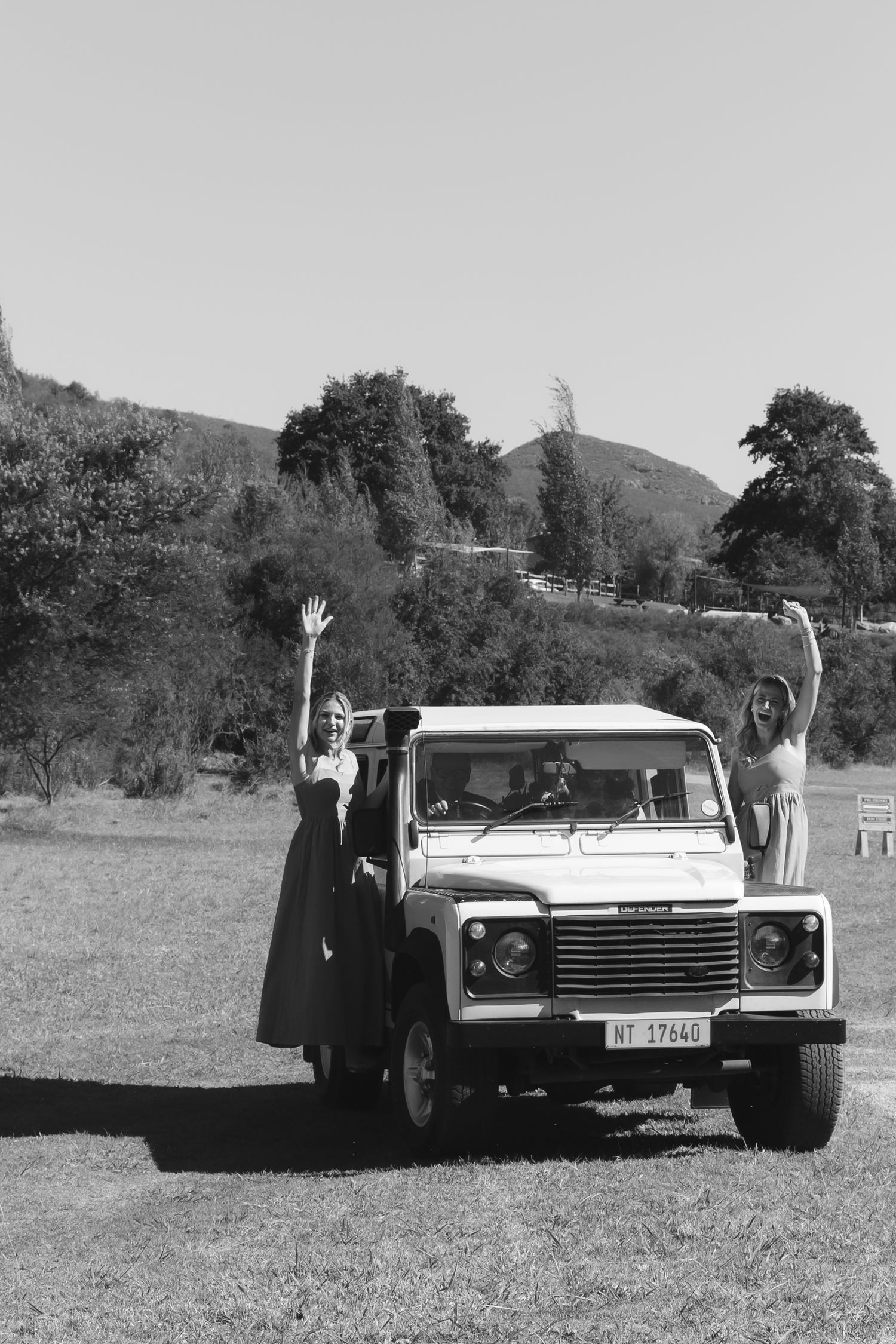Black and white photo series showing vintage cars parked in a field with people waving from beside them.