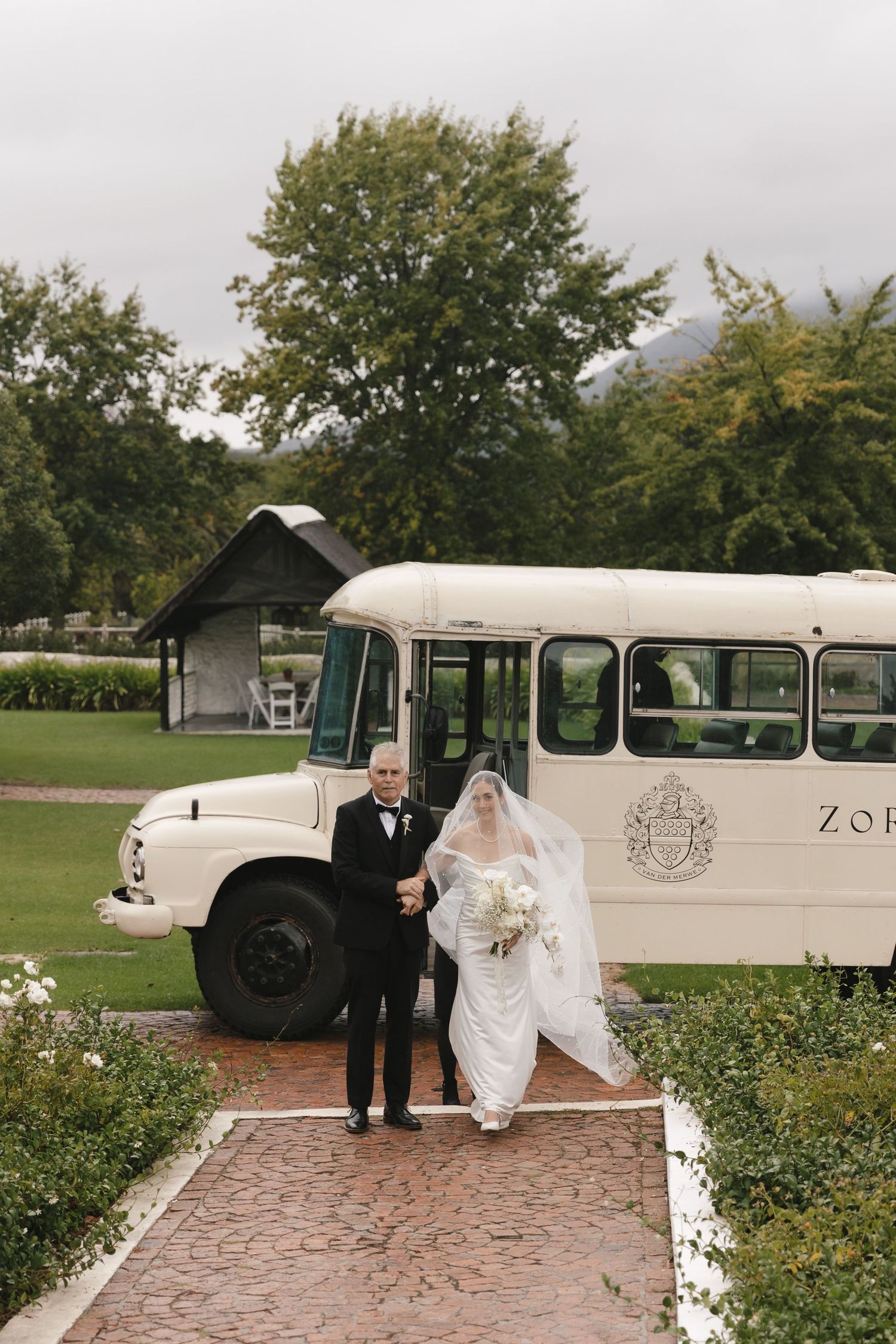 Elegant vintage wedding transportation with classic cream-colored buses parked on brick pavement near garden landscaping.