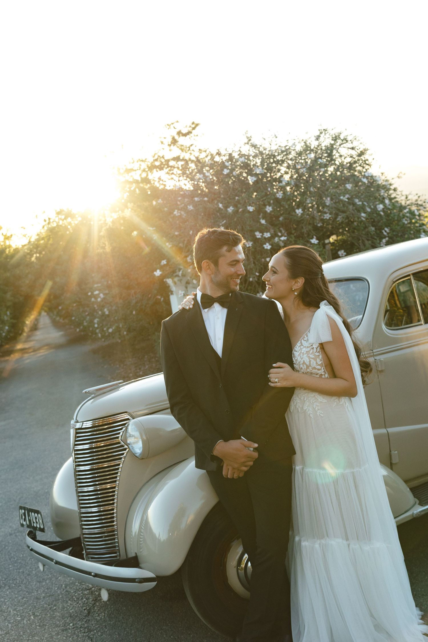 Romantic sunset wedding photos with vintage car featuring flowing white dress and classic styling.