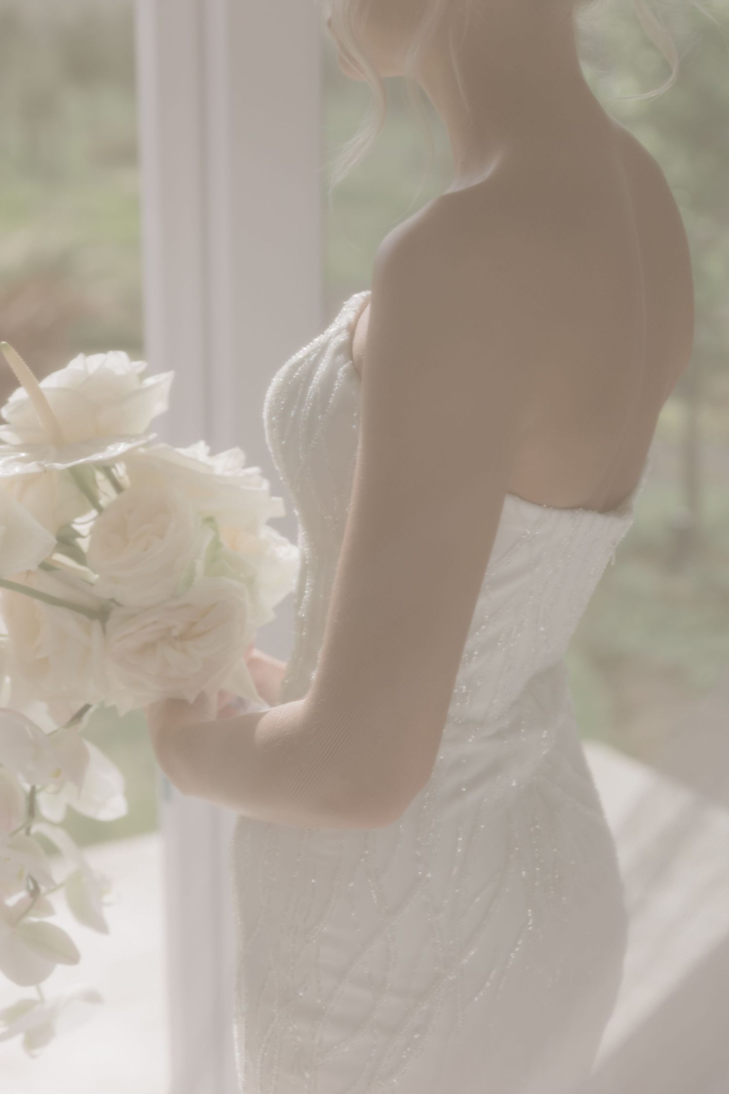 Bride in a beaded strapless wedding gown holding a white floral bouquet near a bright window.