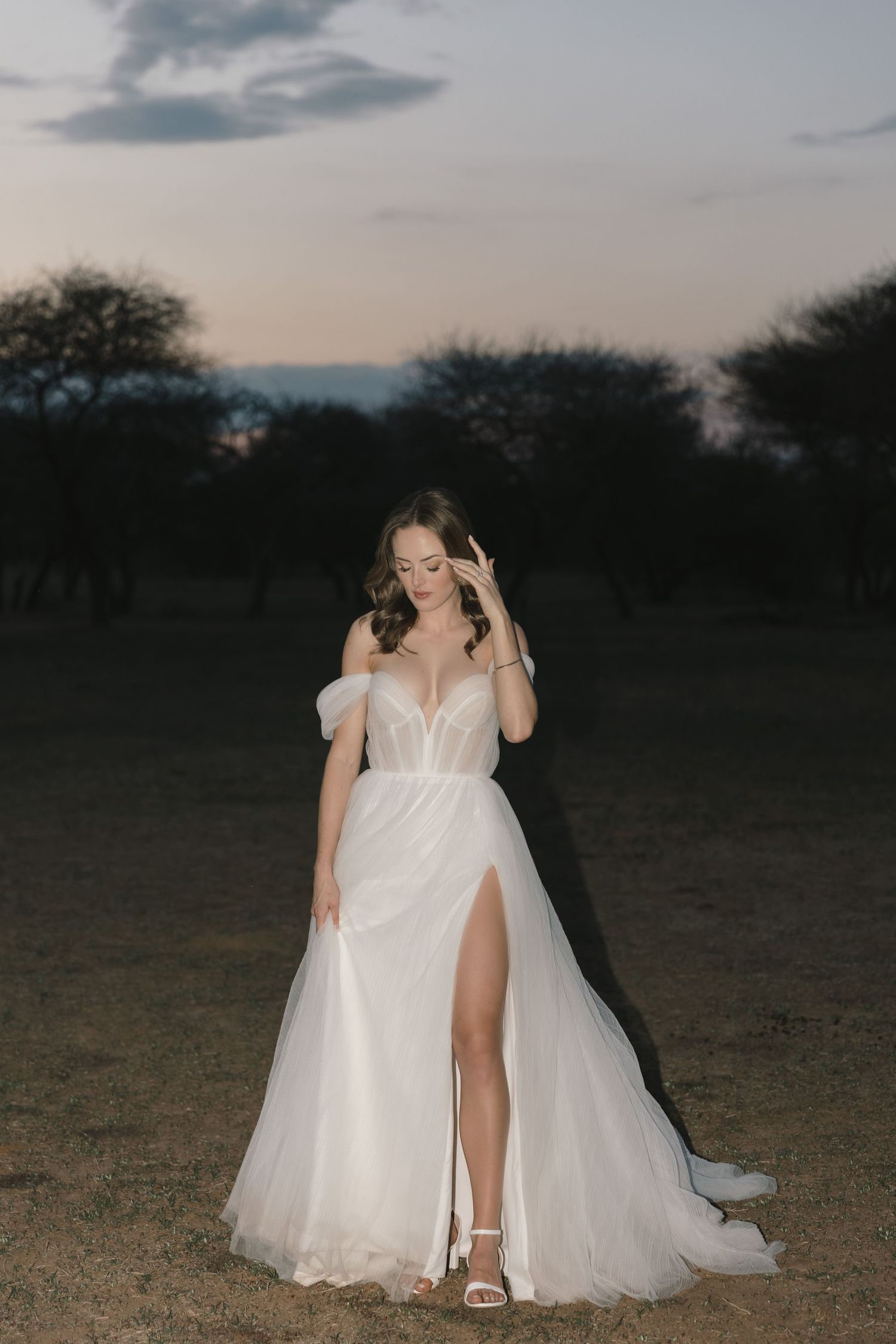 A bride in a flowing white wedding dress sits on the ground at dusk in a natural outdoor setting.