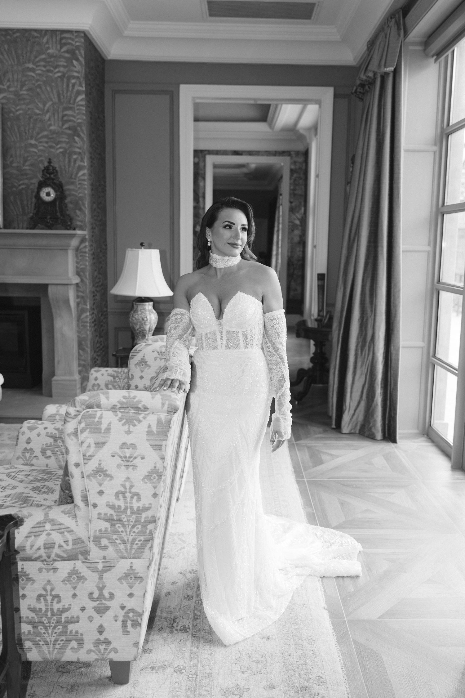 A bride in an elegant white lace wedding dress poses in a bright interior room with damask upholstered chairs.