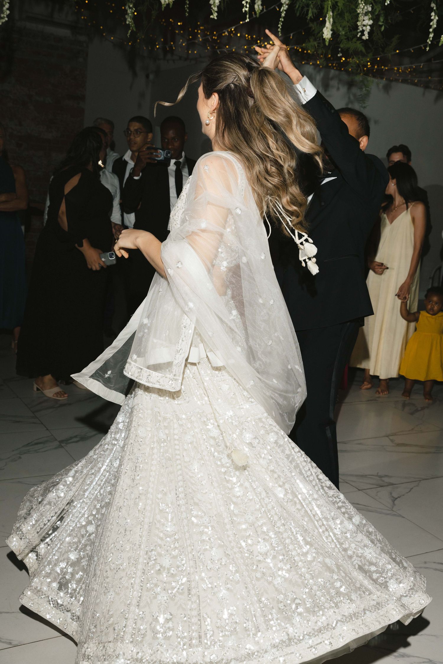 A bride in a sparkling white wedding lehenga twirls on the dance floor during a wedding reception celebration.