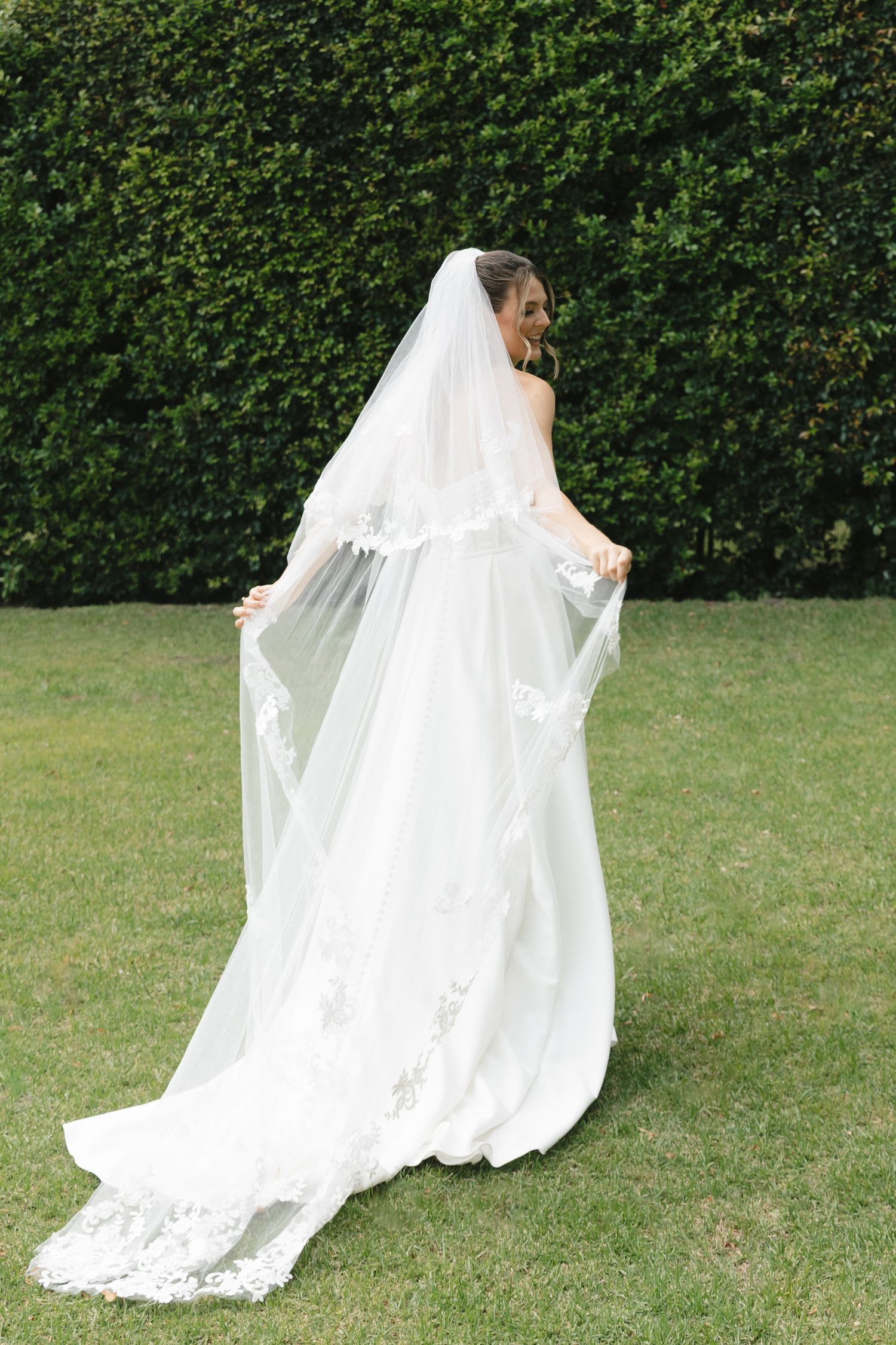 A bride in a flowing white wedding dress and veil twirls gracefully on a green lawn against a hedge backdrop.