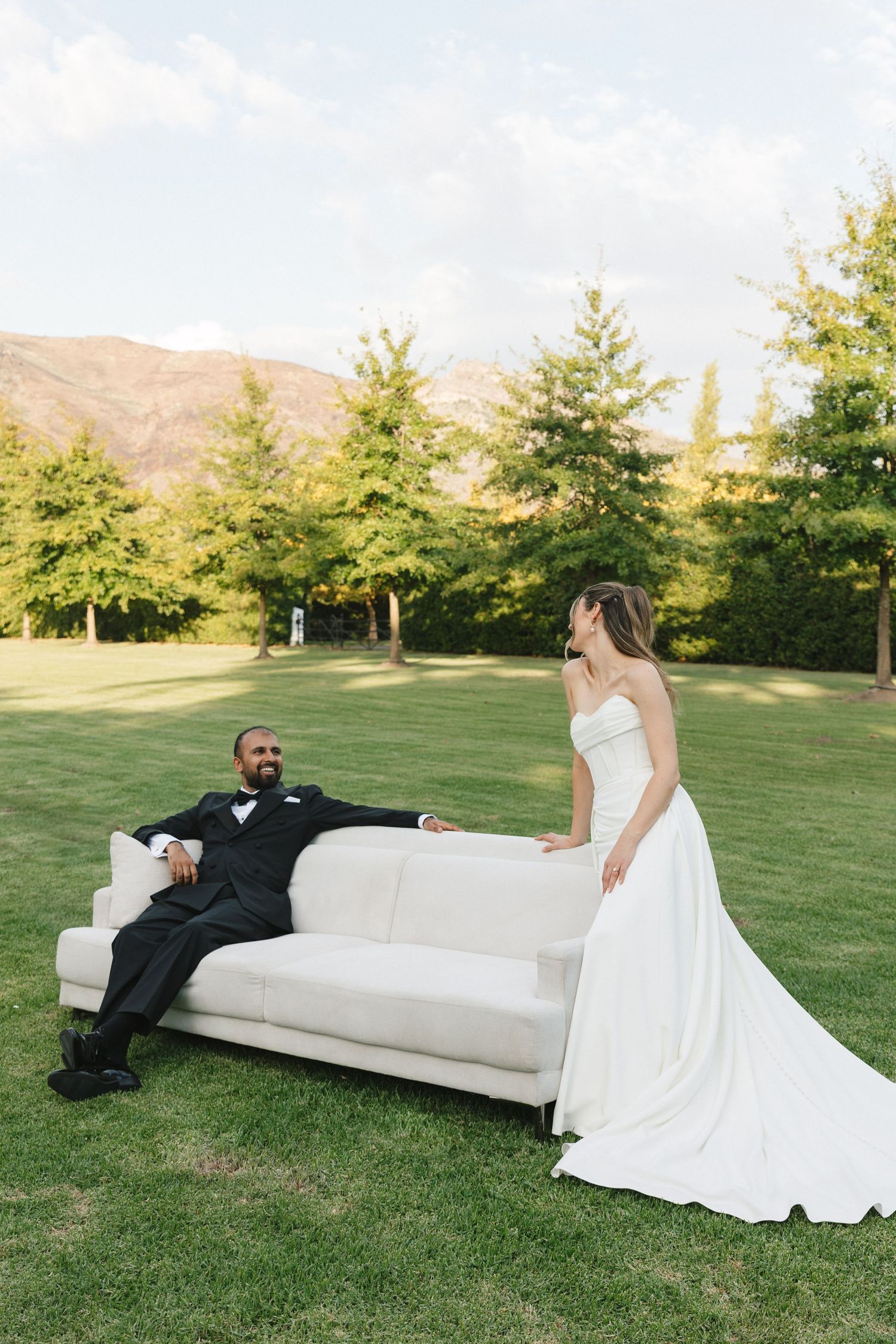 Couple relaxes on white outdoor couch during wedding photoshoot on grassy hillside.