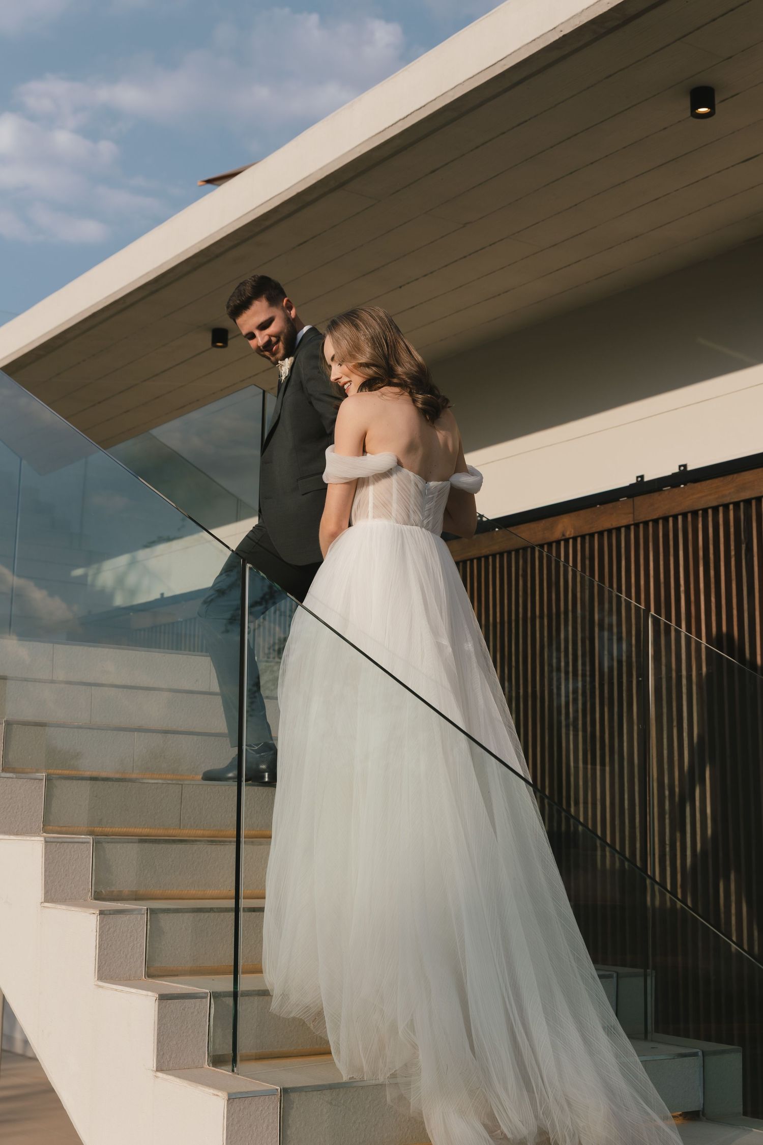 A couple shares a romantic embrace on a modern balcony with white architectural details and glass railings at sunset.