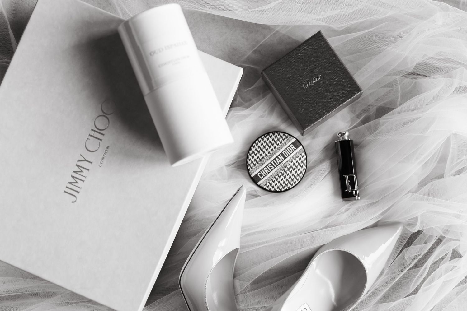 Black and white flat lay of Jimmy Choo box, Cartier compact, lipstick, and heels on tulle fabric.