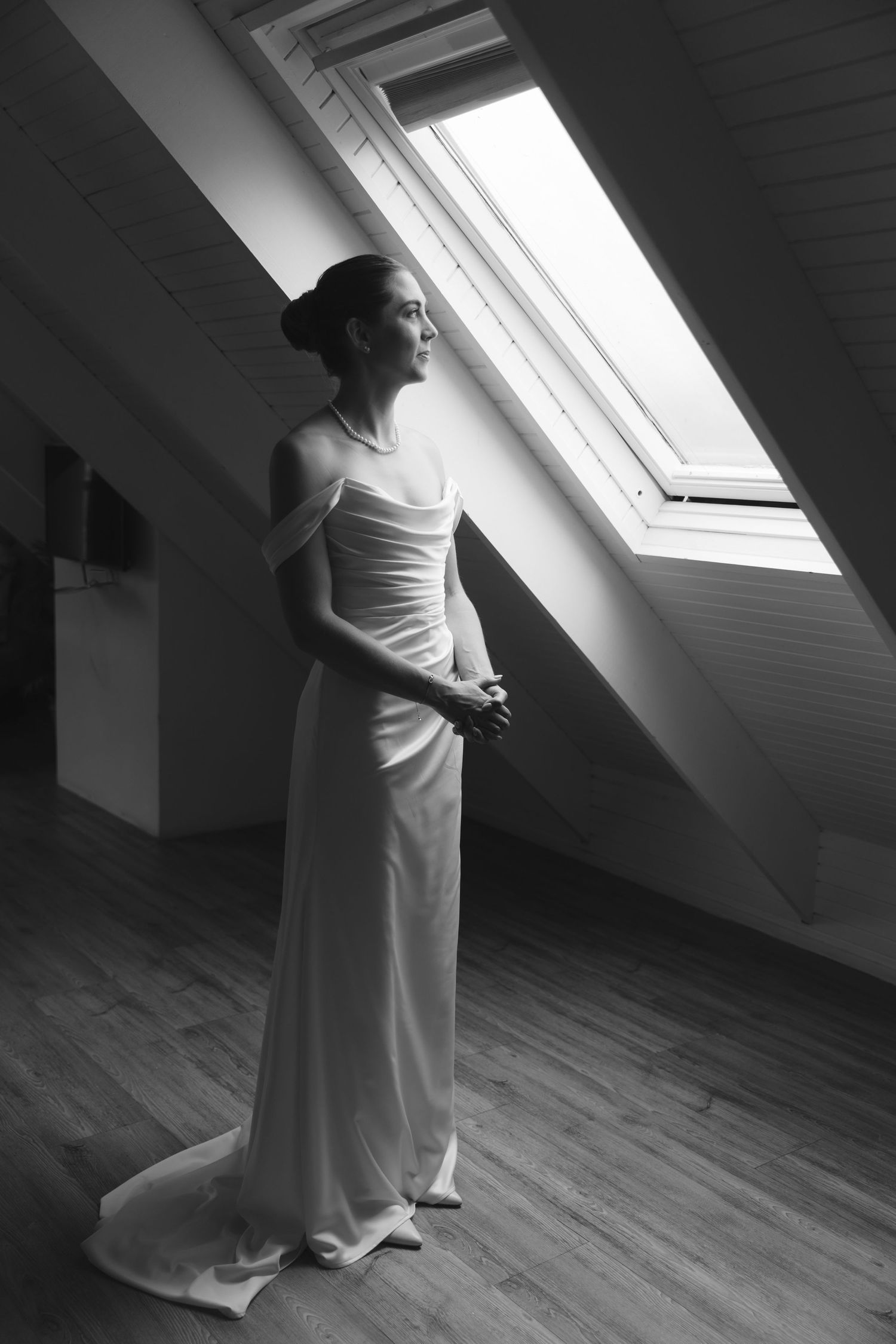 A figure in a flowing white dress stands in dramatic lighting beneath a slanted skylight window in a minimalist attic space.