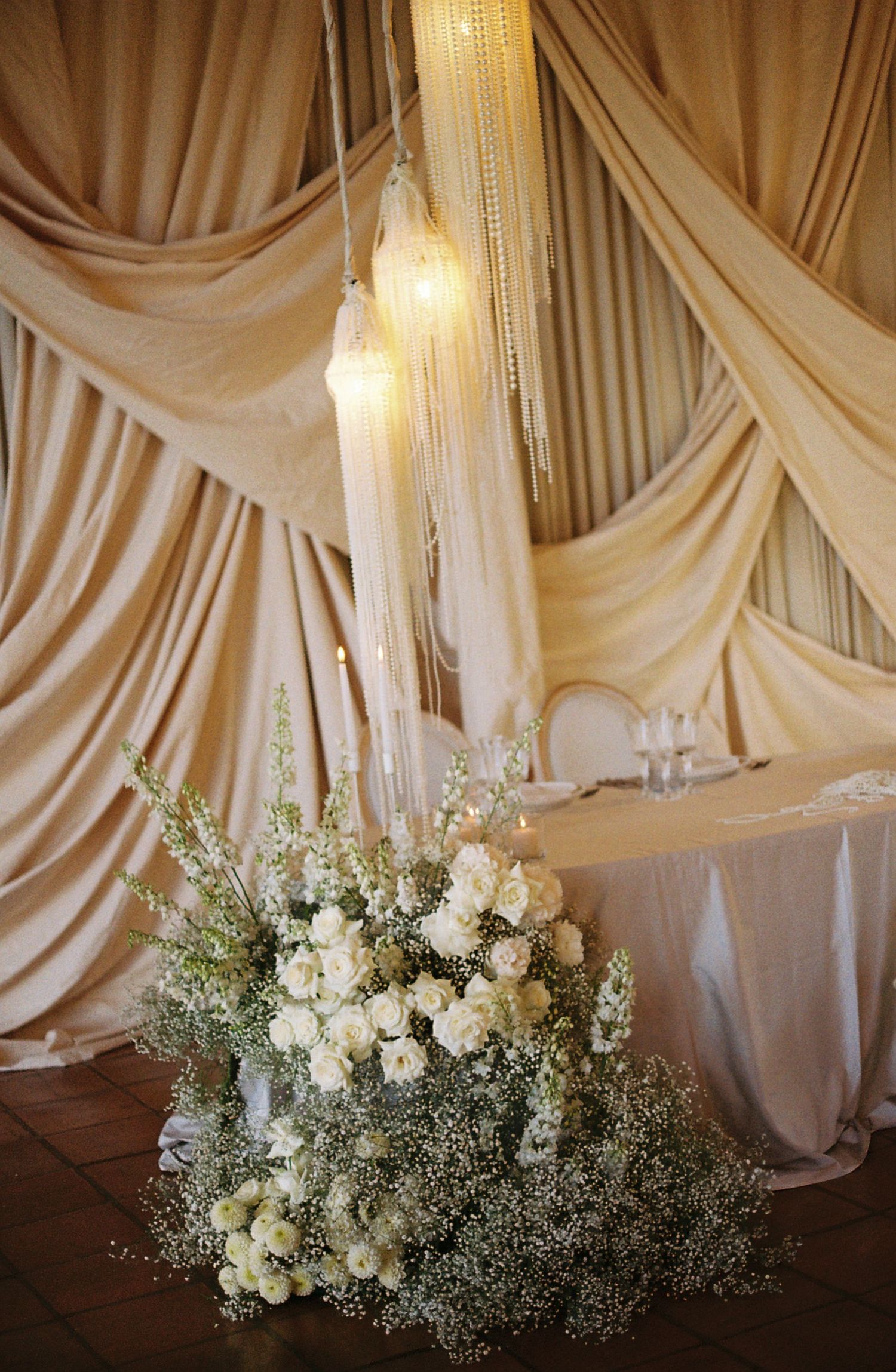 White floral arrangement with baby's breath under elegant draped fabric and hanging lights.