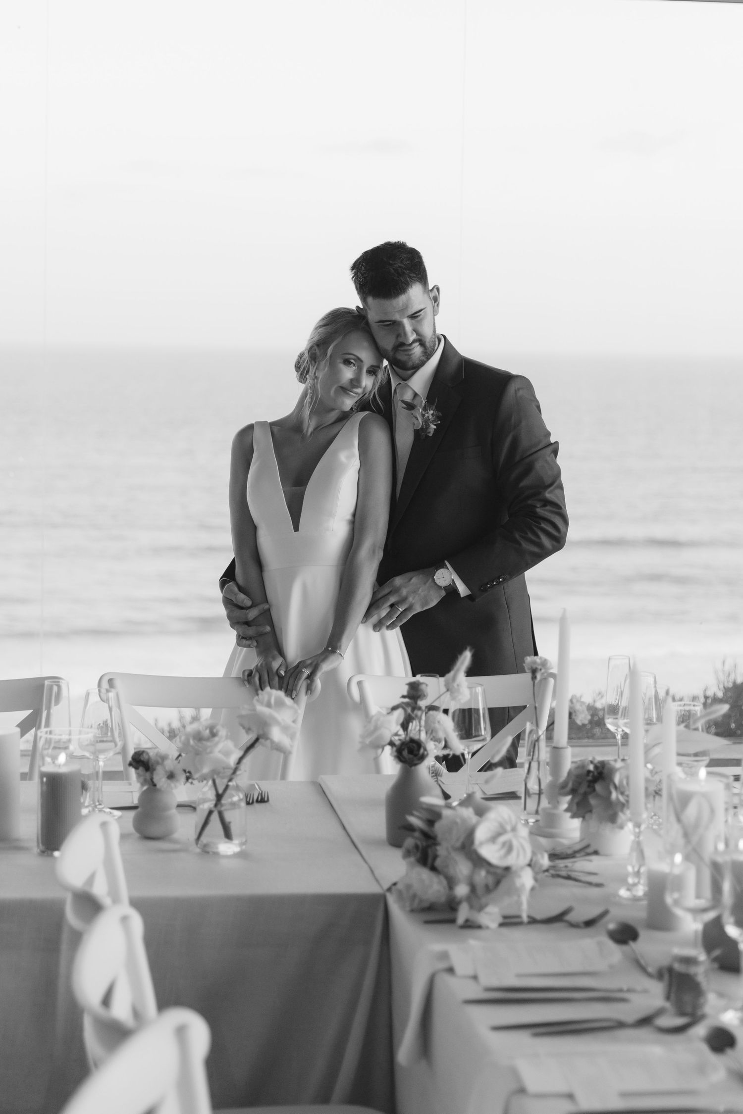 A couple shares an intimate moment at an elegant seaside wedding reception with white table settings and floral centerpieces.