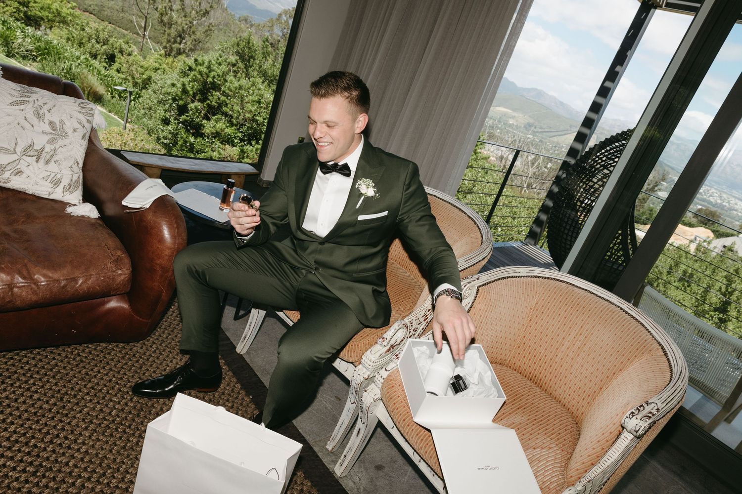 Groom in green tuxedo holding drink, opening gifts in elegant room with mountain views.