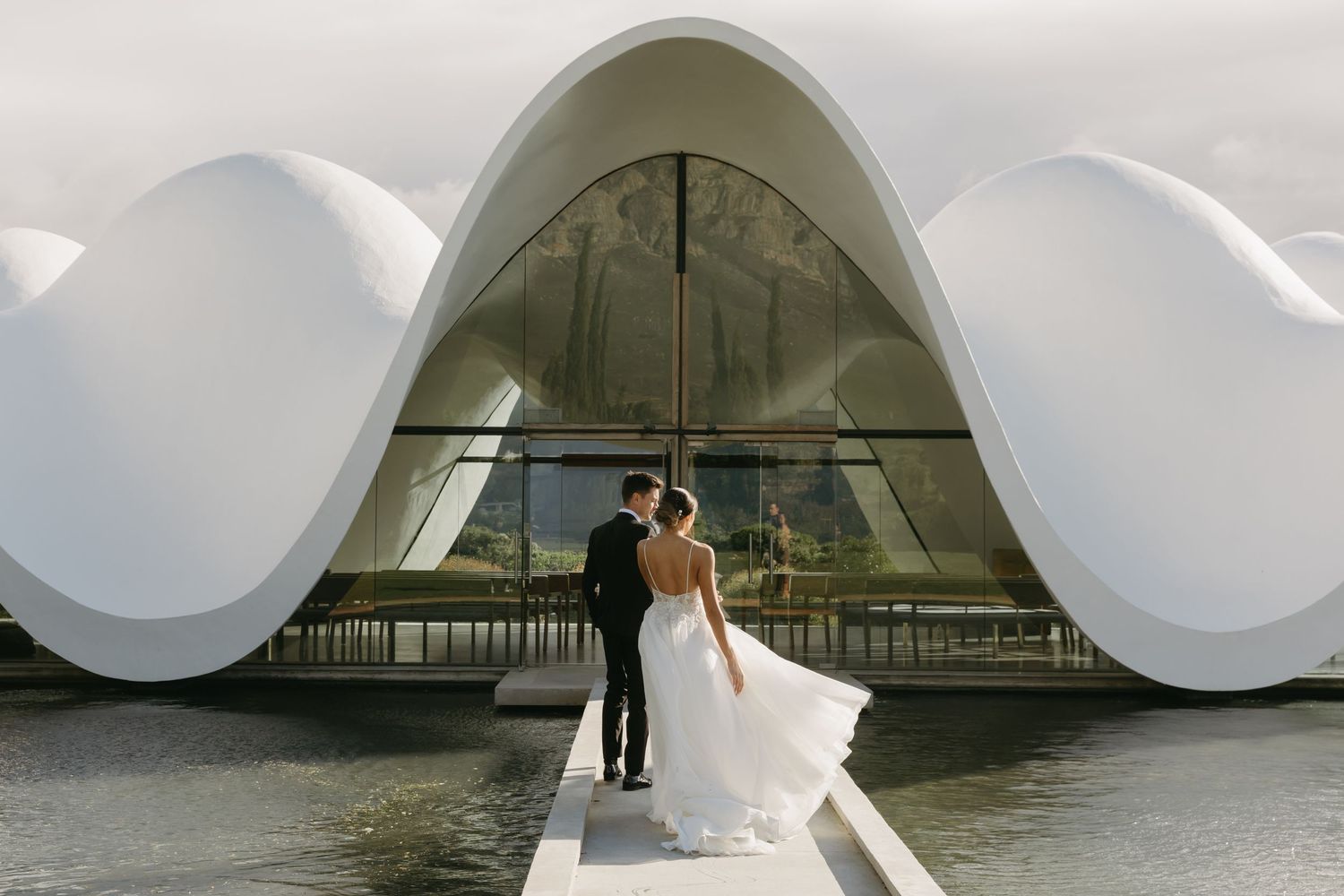 Bride and groom stand on a dock before a stunning white curved modern wedding venue reflected in calm water.