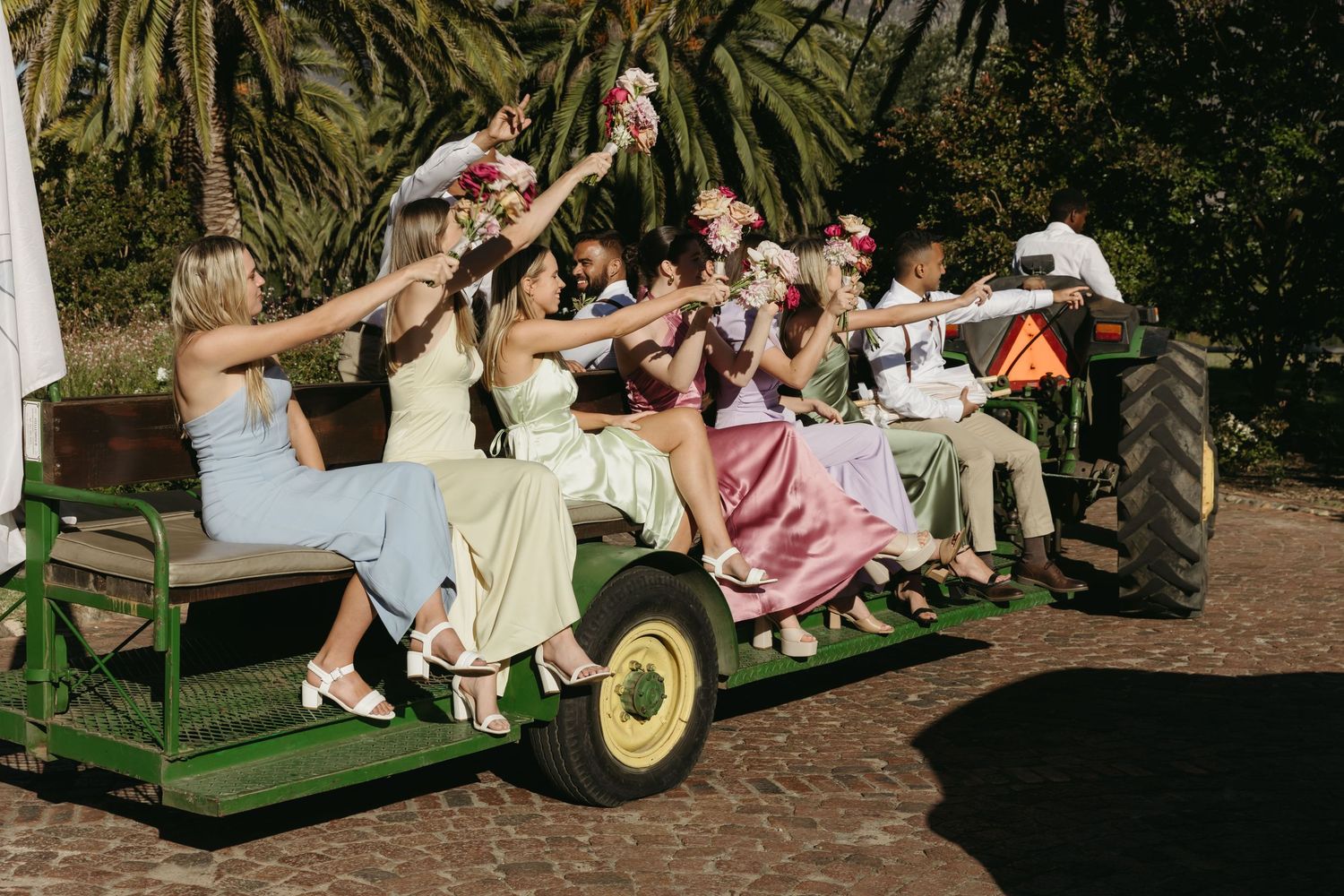 Wedding party in colorful dresses riding a green tractor trailer, celebrating outdoors near palm trees.