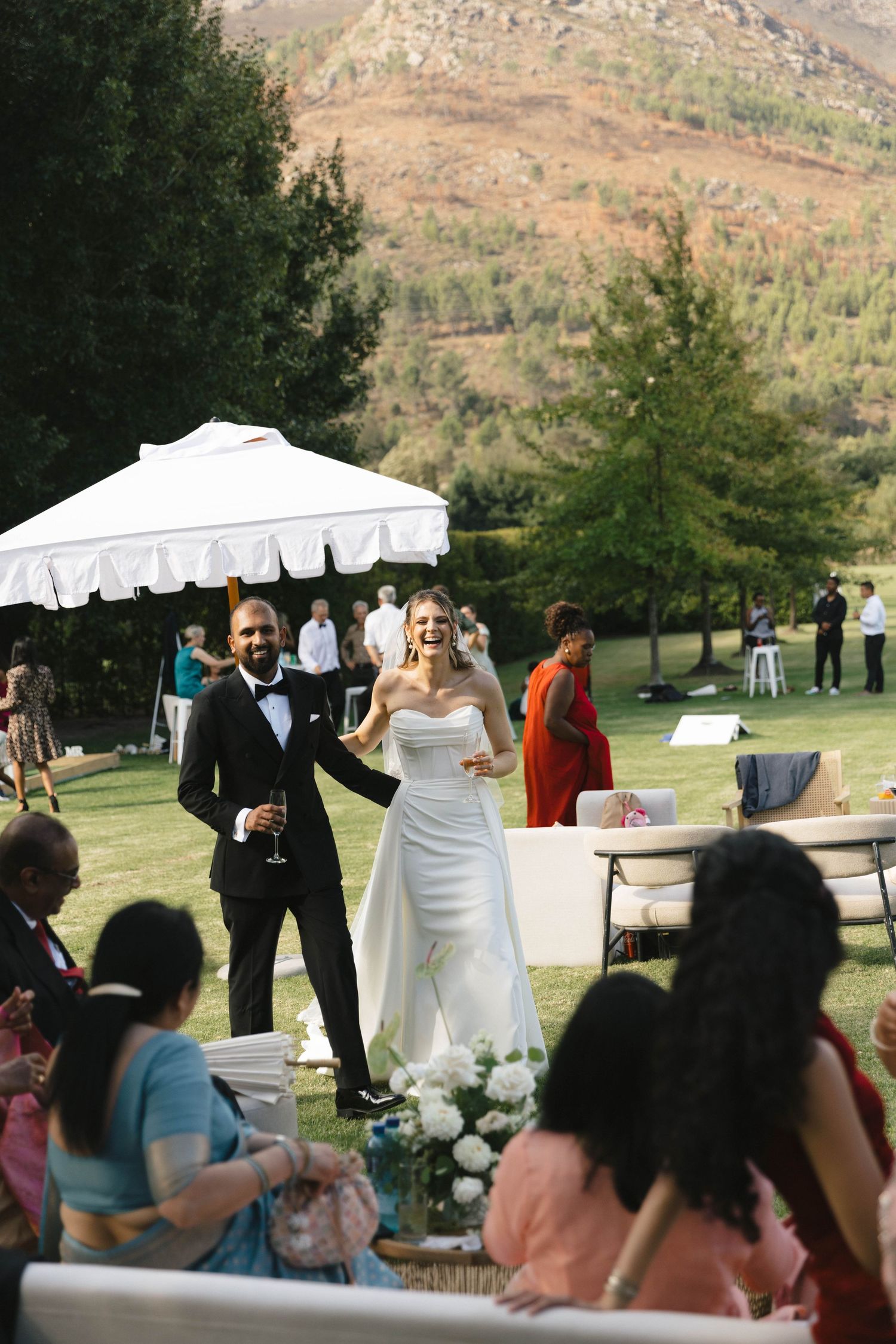 A wedding celebration takes place outdoors with a white tent against a scenic mountain backdrop in natural lighting.