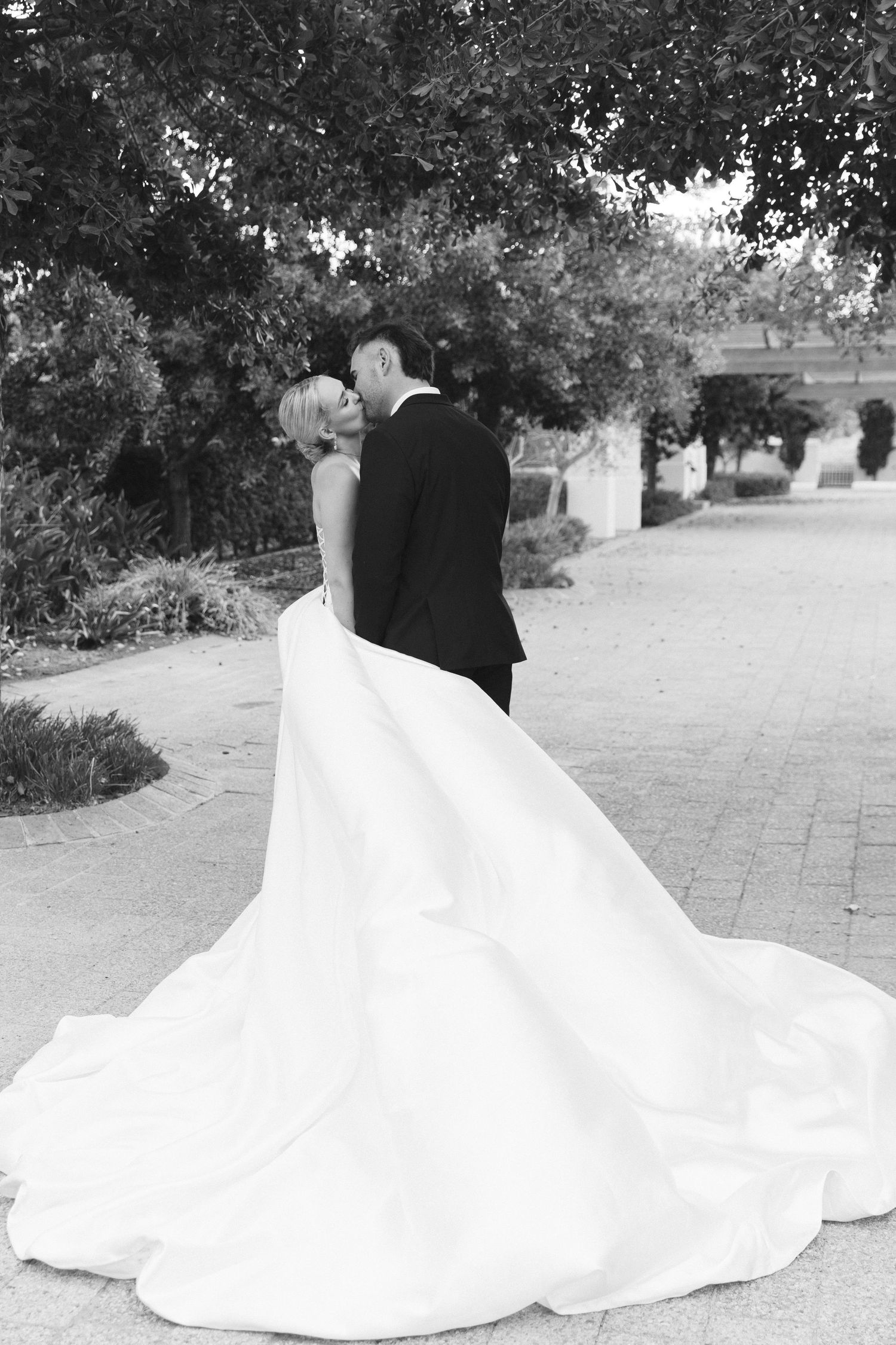 A flowing white wedding dress train spreads elegantly across stone pavement in a black and white outdoor photo.