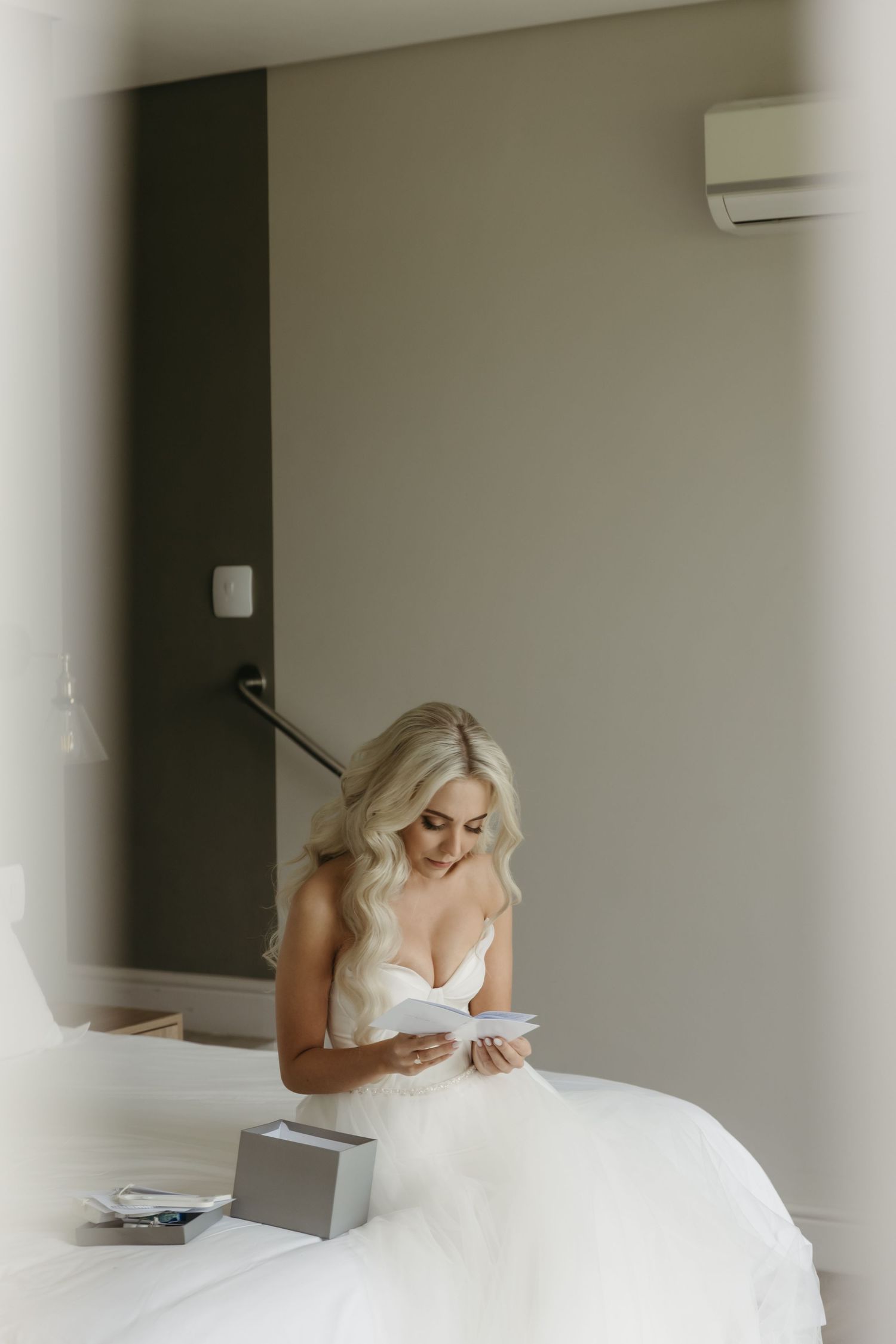Blonde bride in white wedding dress sits on bed reading a heartfelt letter in a modern hotel room.