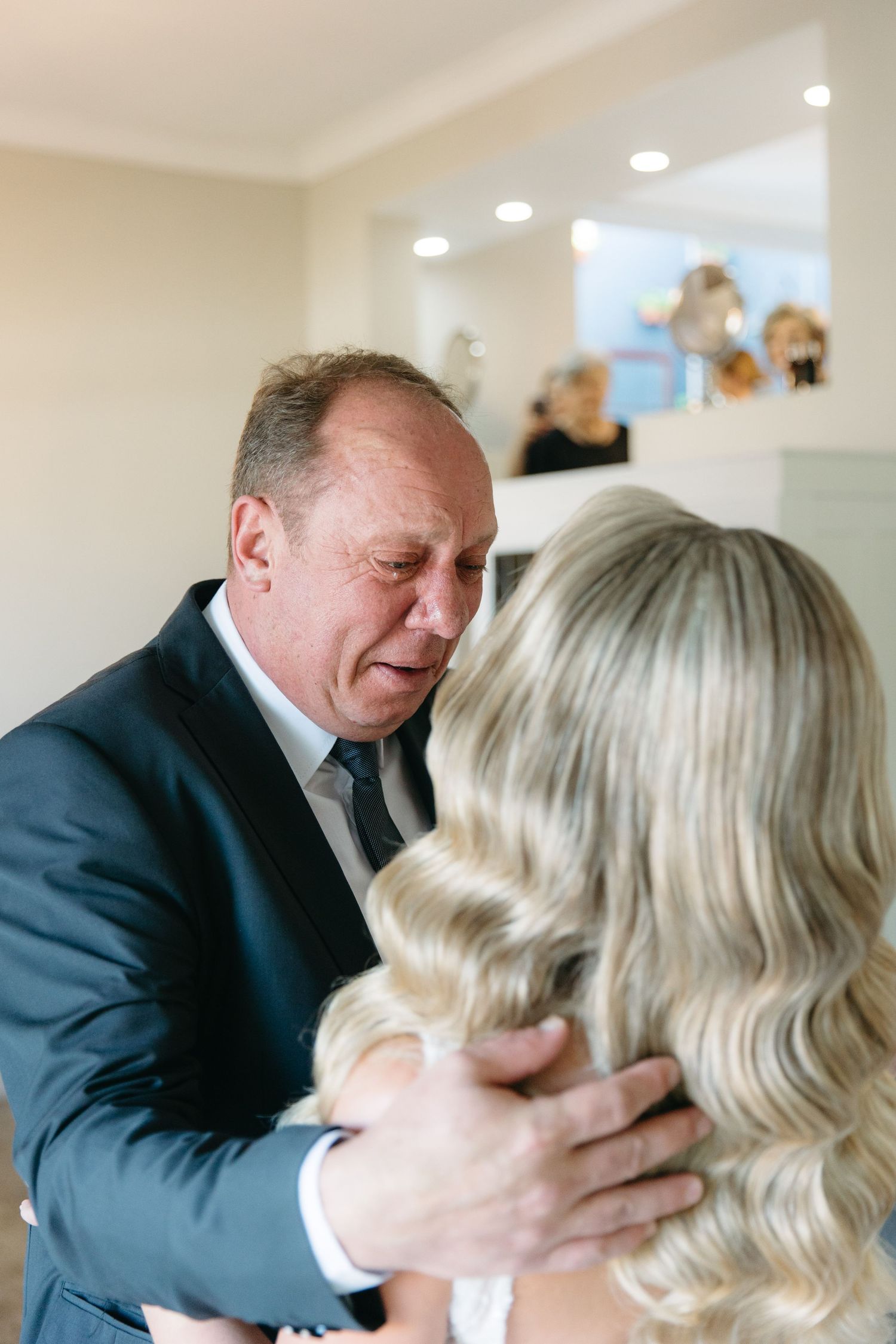 Emotional father embracing bride in white dress during wedding day first look moment indoors.