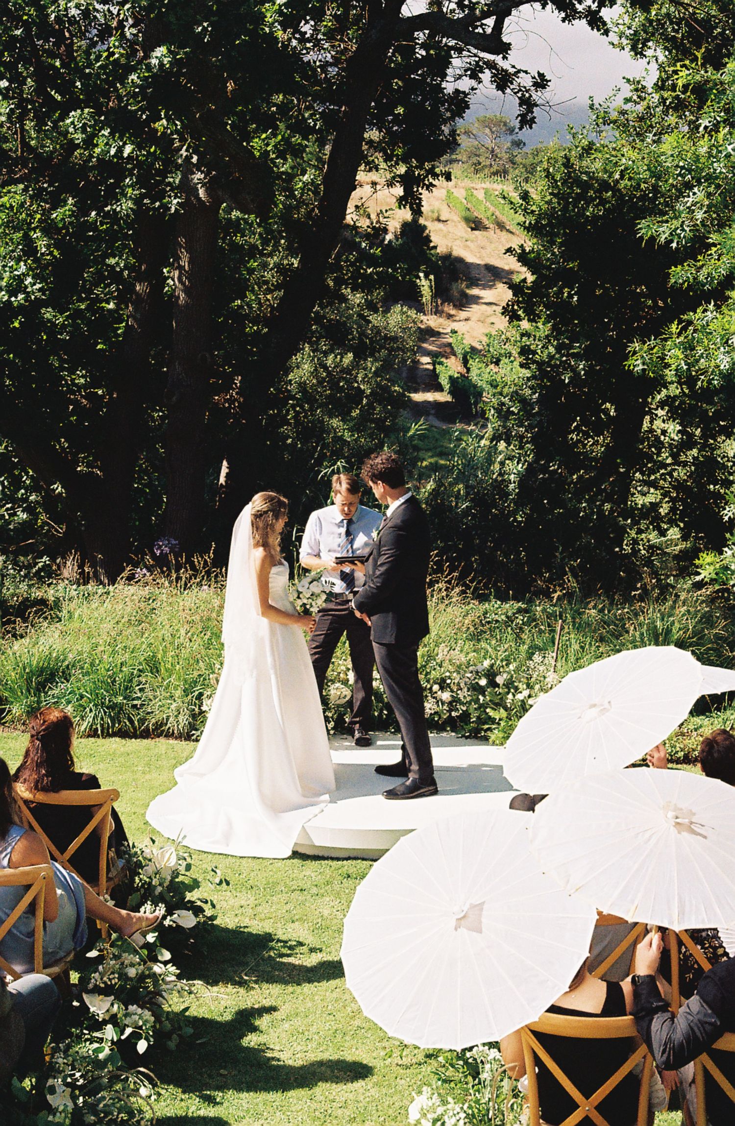 Outdoor wedding ceremony setup with wooden chairs and white aisle runner in a garden setting.