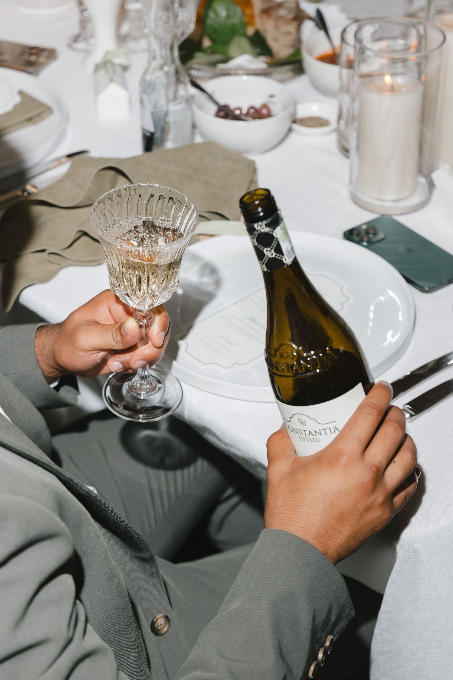 Hands holding wine glasses and bottles at an elegant table setting with white linens and candlelight.