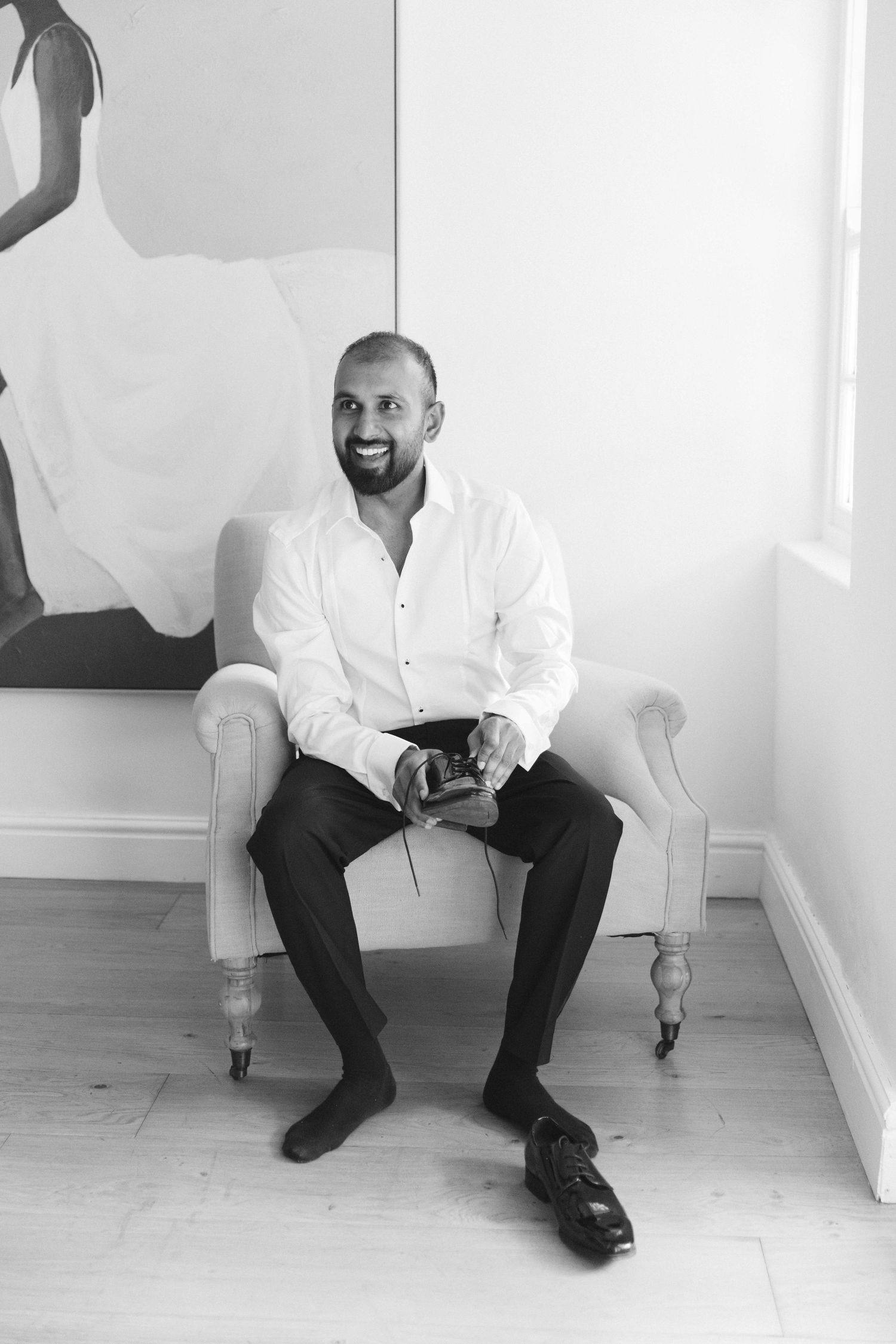 A person in a white shirt and dark pants sits casually on the floor against a white wall in a minimalist setting.