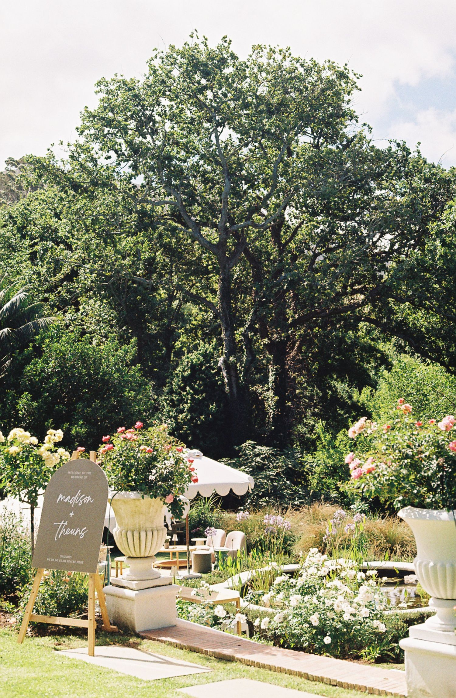 Elegant outdoor garden wedding venue with white seating and floral arrangements along a stone pathway under shady trees.