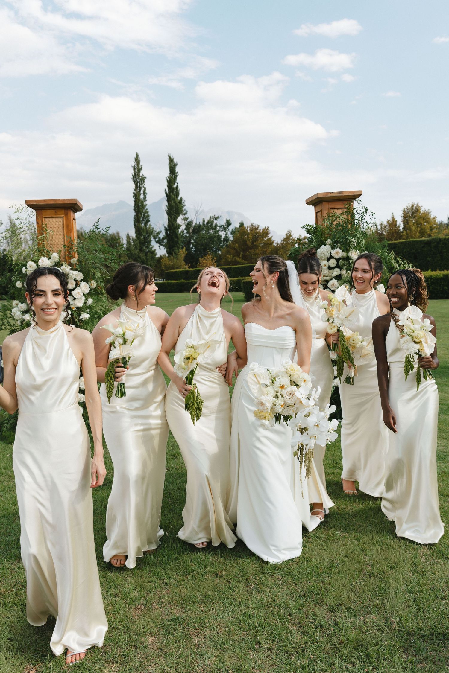 Bridesmaids in elegant white dresses holding bouquets laugh together during outdoor wedding photoshoot.