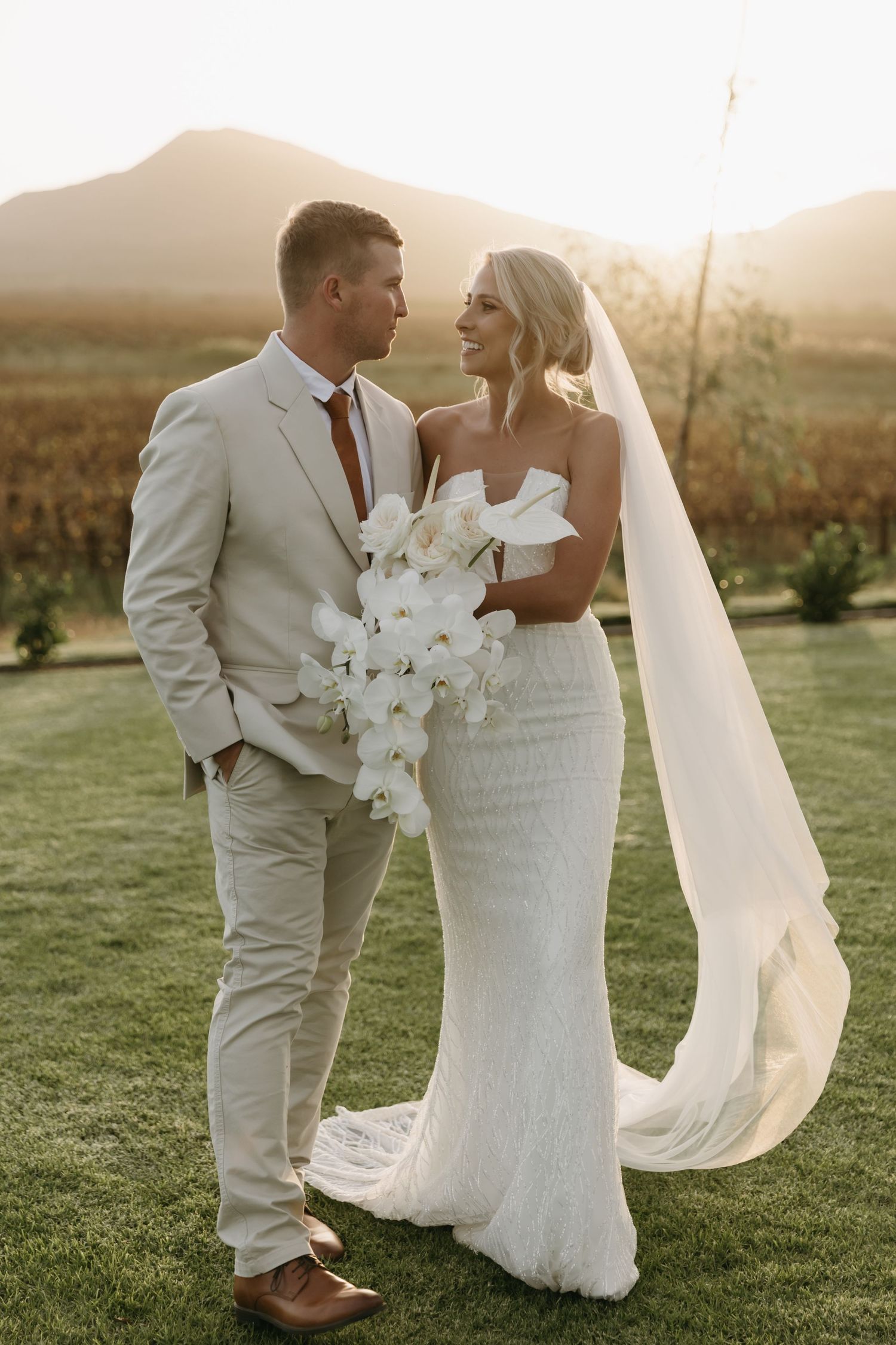 Bride and groom share a romantic moment at sunset in a vineyard, she in a white gown with veil, he in a beige suit.