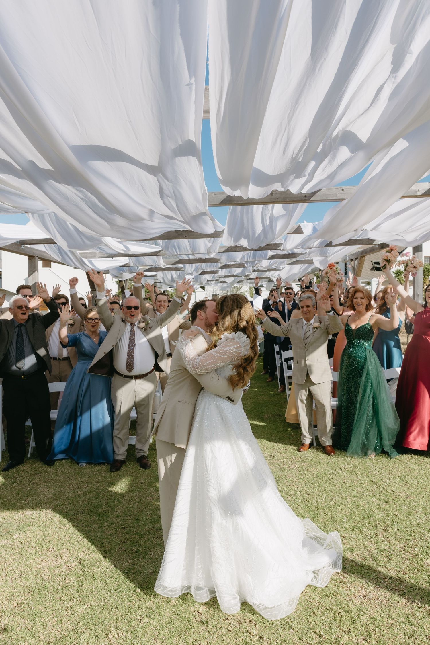 Bride and groom share first kiss at outdoor wedding ceremony under white fabric canopy with cheering guests.