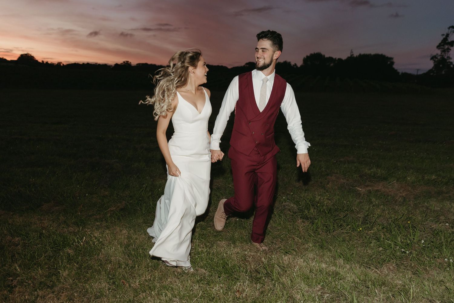 A couple in wedding attire walks through a field at sunset holding hands while their photographer captures candid moments.