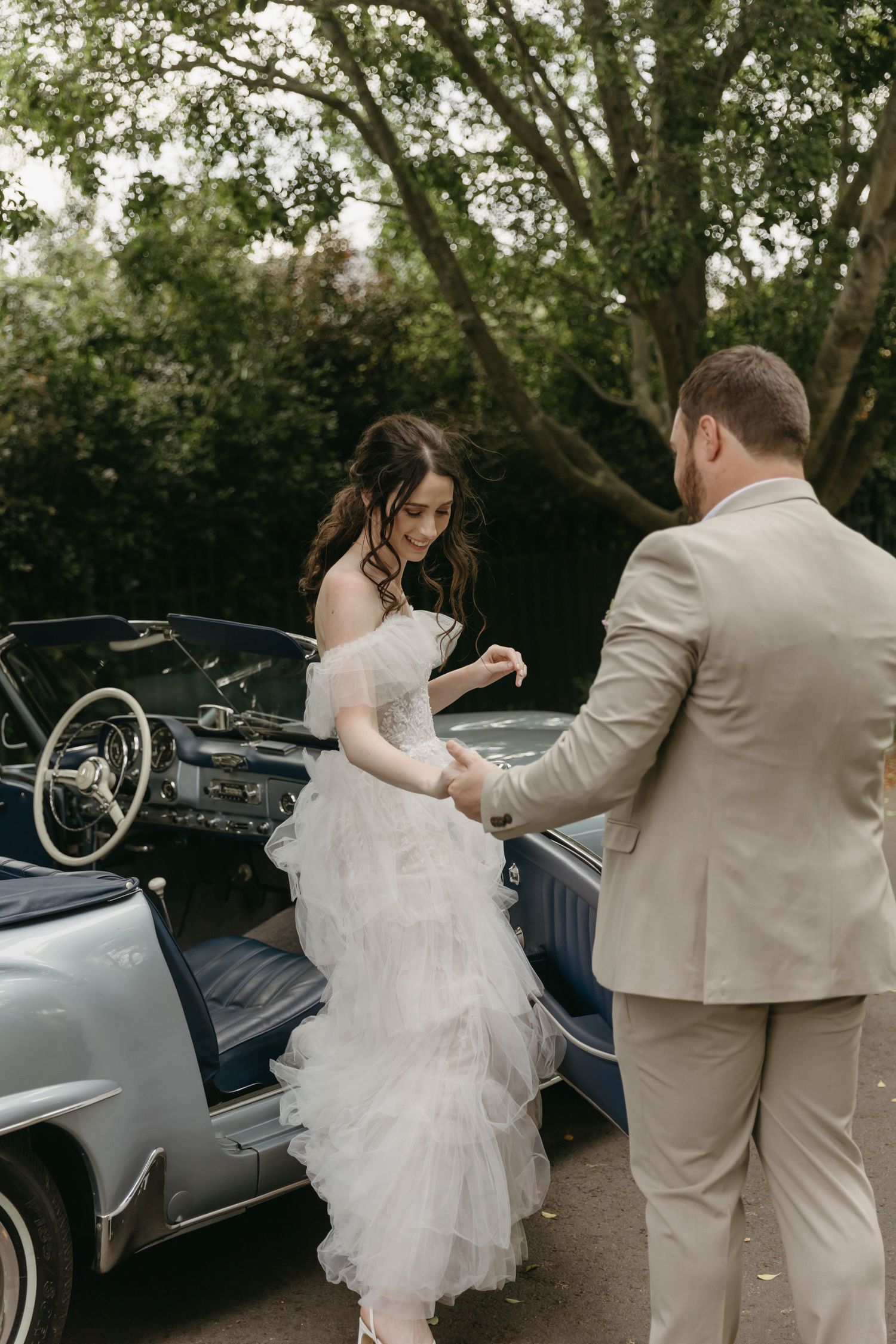 Groom in beige suit helps bride in white tulle gown exit a vintage blue convertible car outdoors.