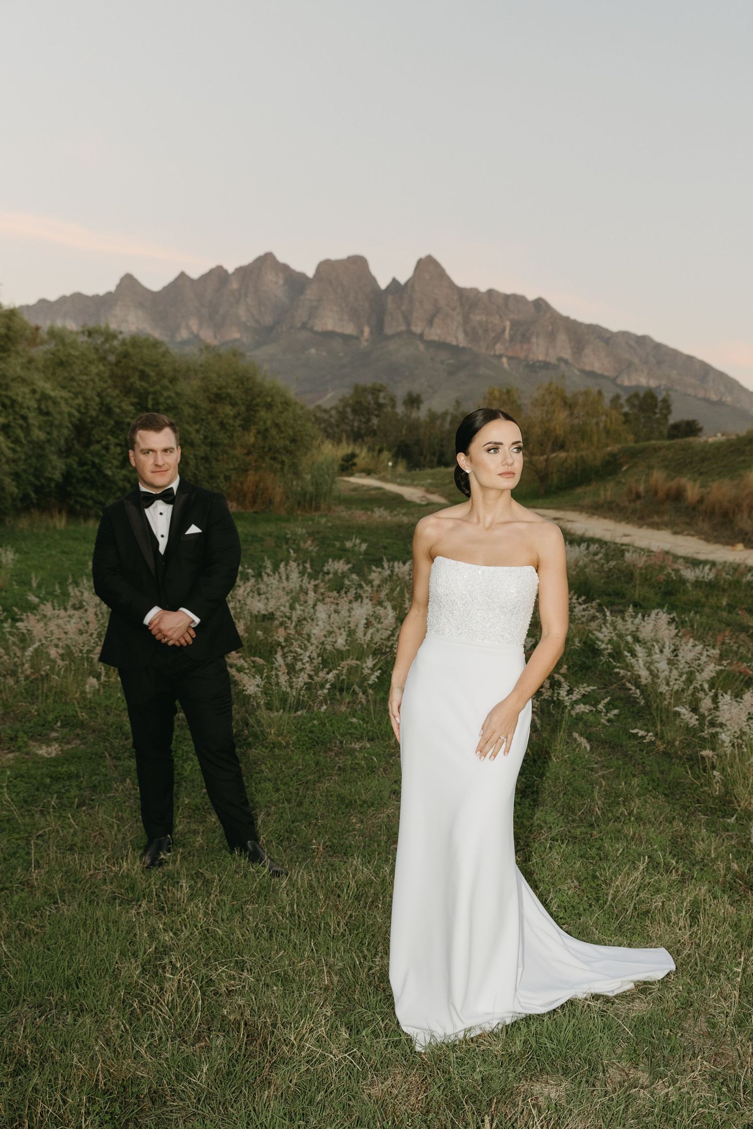 Bride in white strapless gown and groom in black tuxedo pose before dramatic mountain backdrop at sunset.