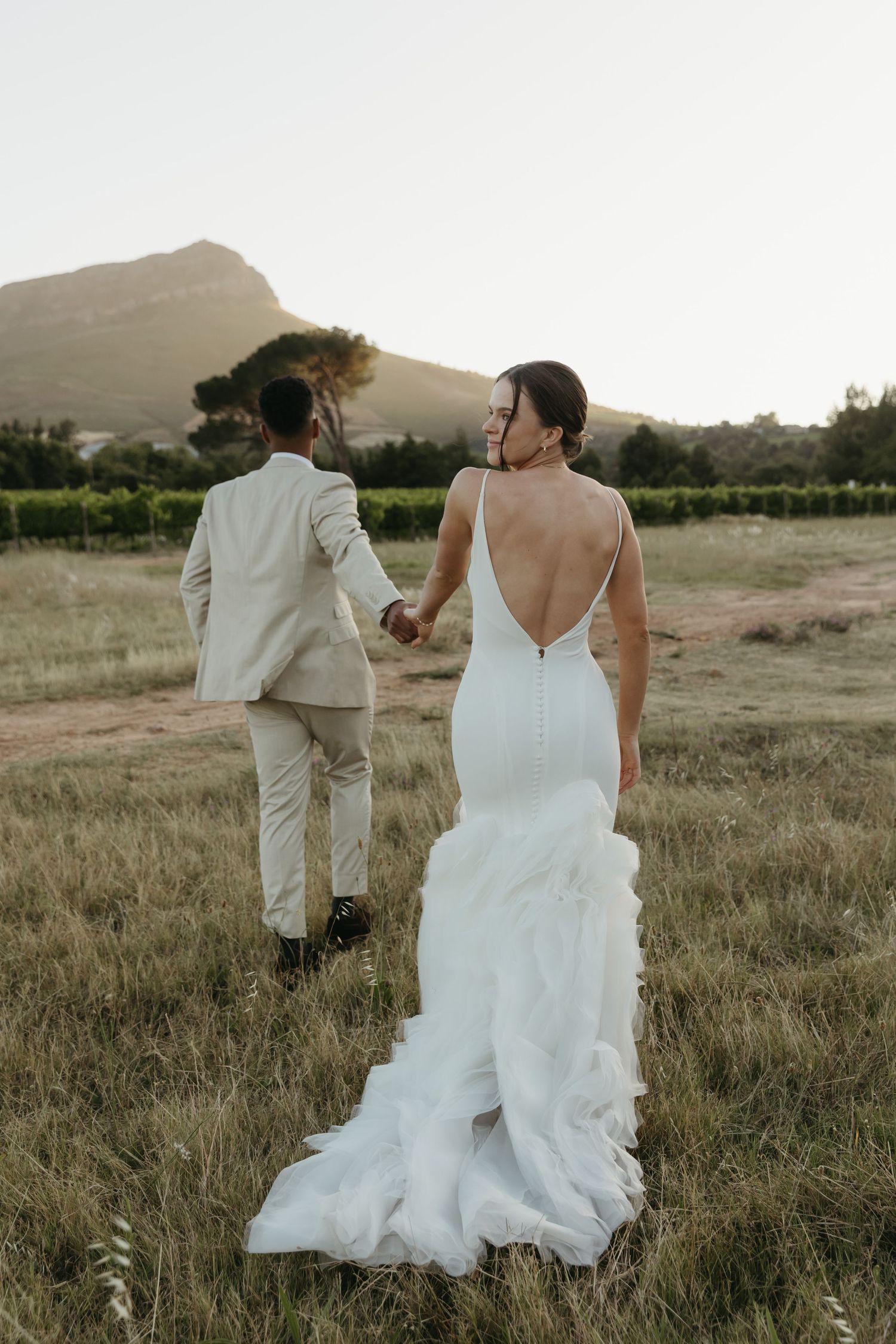 Bride in backless white gown holds groom's hand walking through vineyard field with mountain backdrop at sunset.