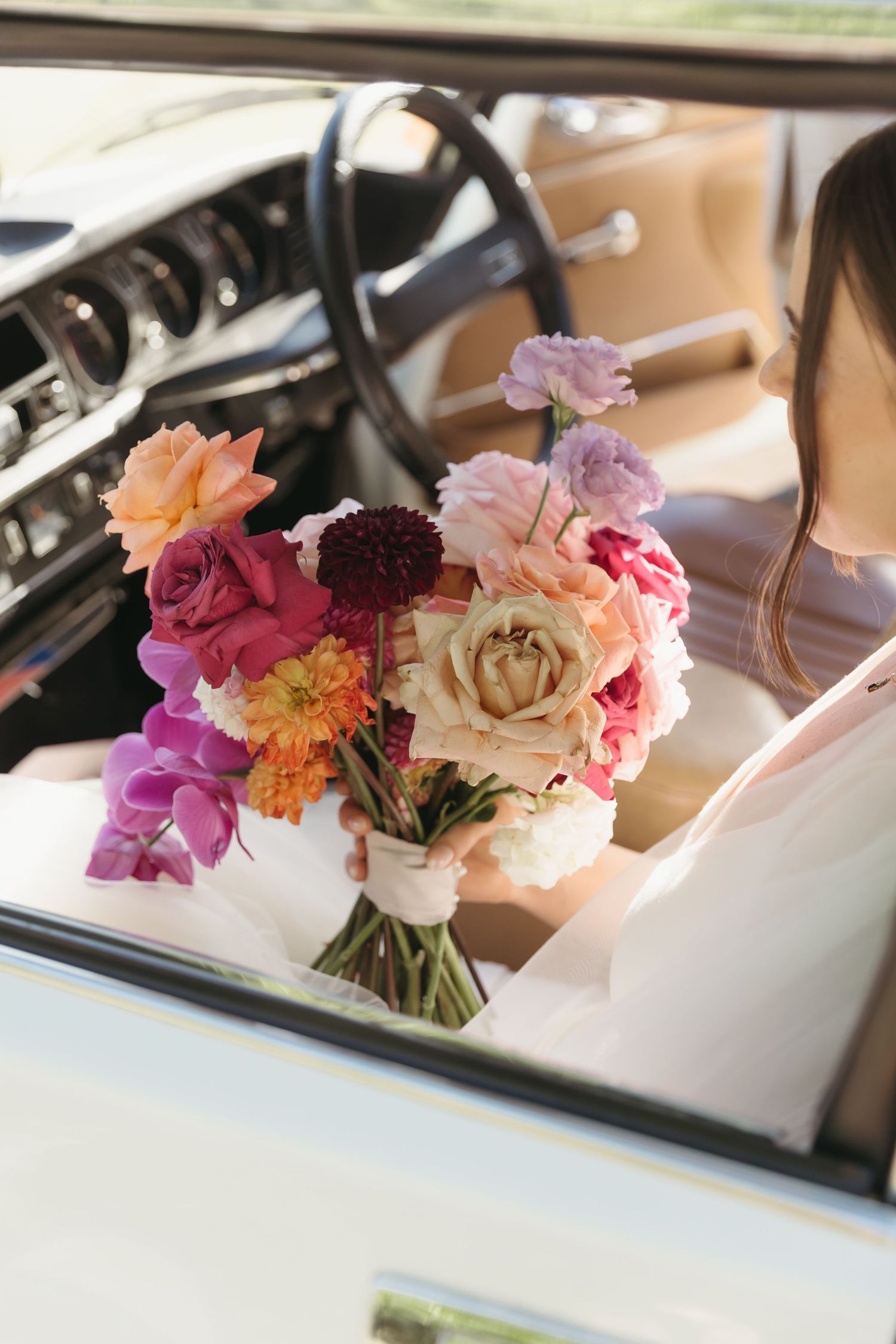 Bride in white dress holding colorful wedding bouquet with roses and dahlias while sitting in a vintage classic car.