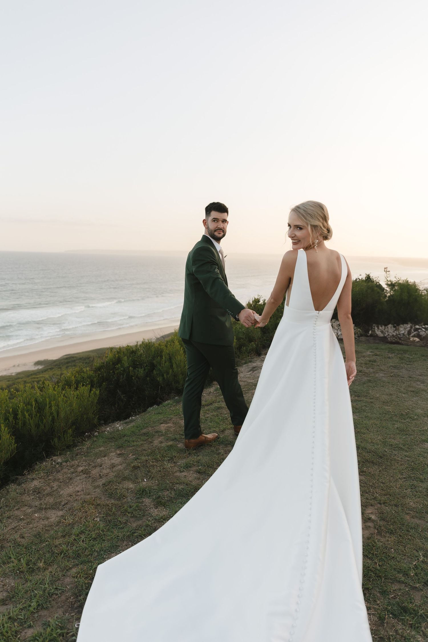 A couple in wedding attire walks along a scenic coastal cliff at sunset with flowing white dress in the ocean breeze.
