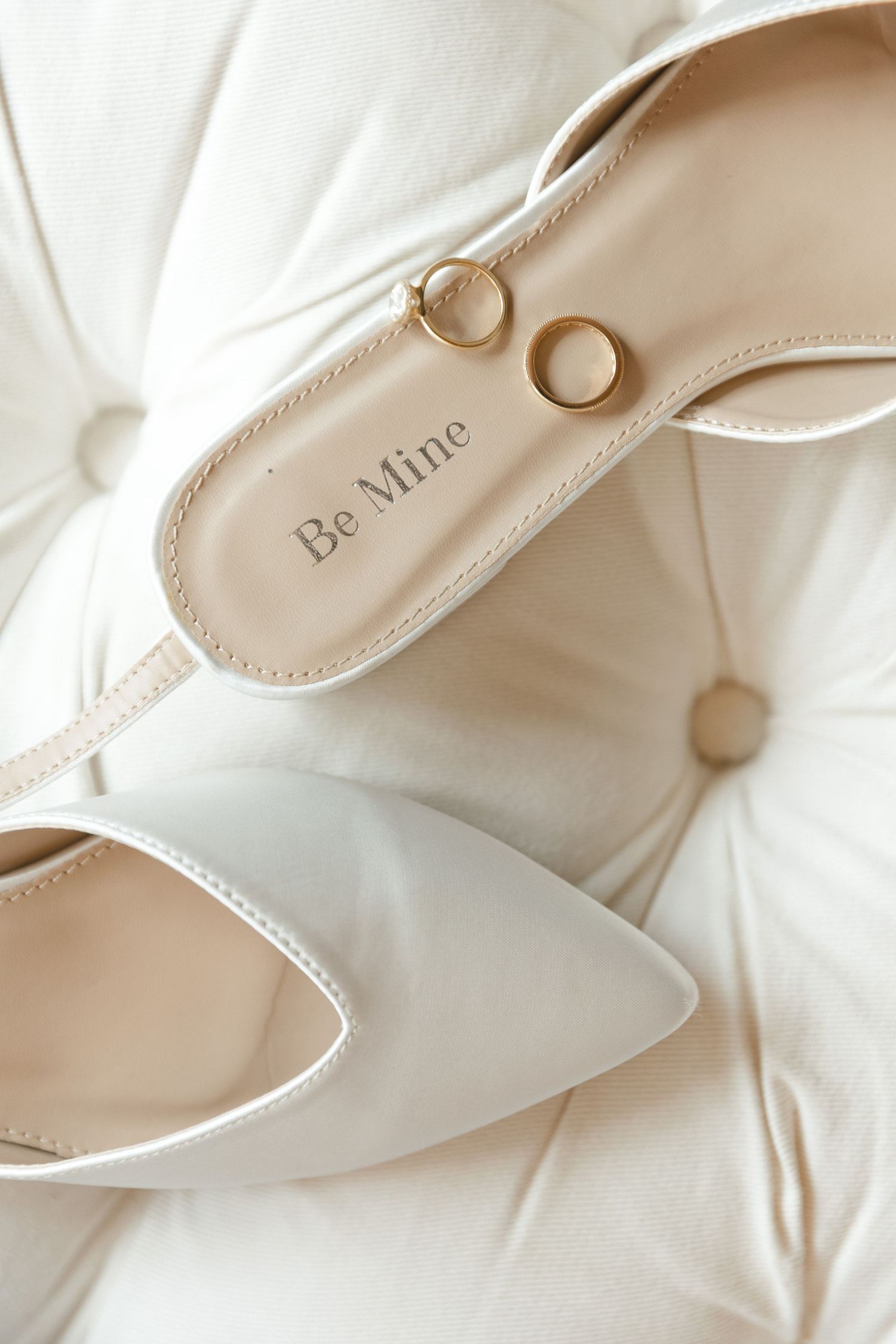 Wedding rings rest on a white bridal shoe with 'Be Mine' text inscribed on the sole against a soft white background.