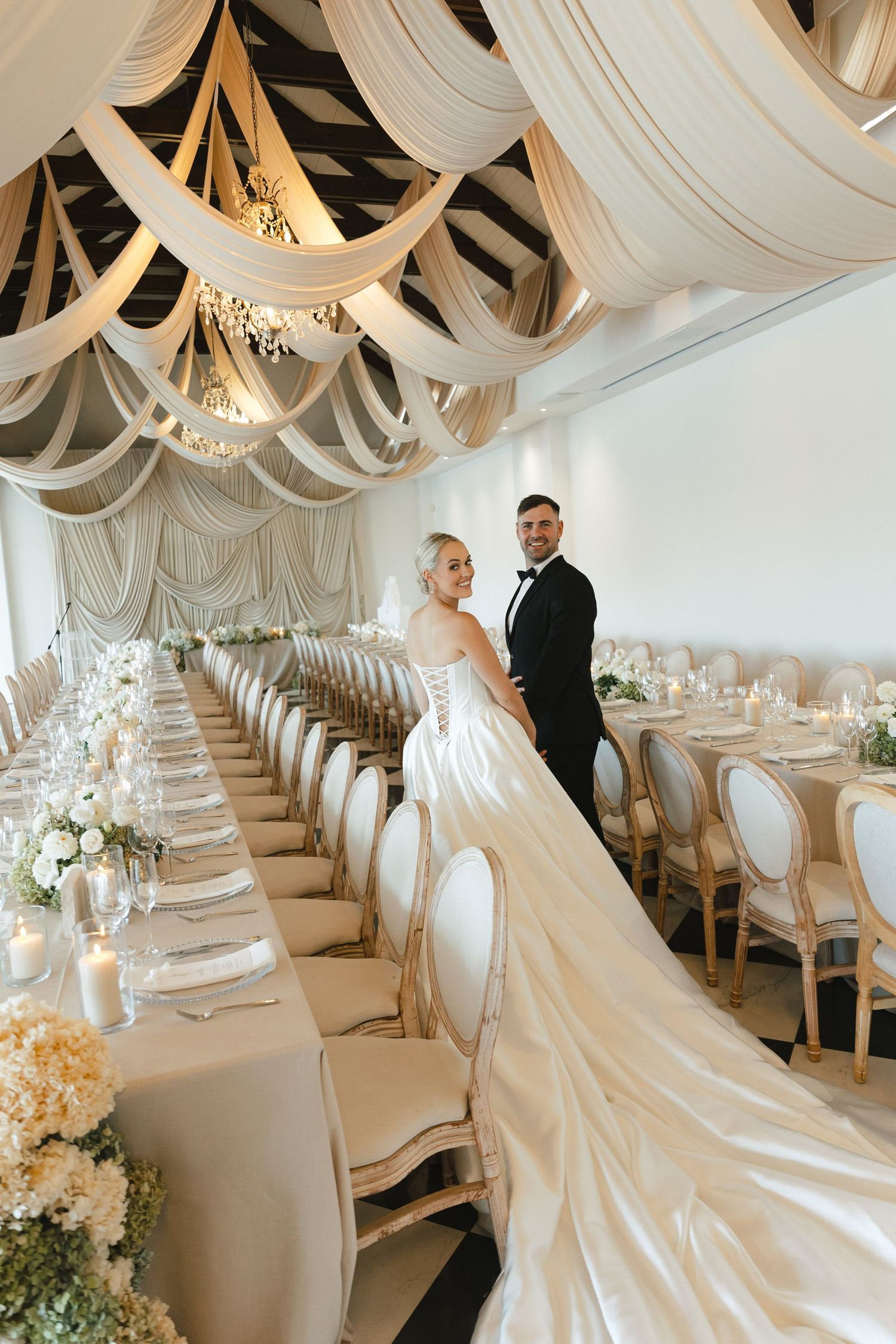 Elegant wedding reception venue with draped white fabric ceiling, crystal chandeliers, and long dining tables set in white decor.