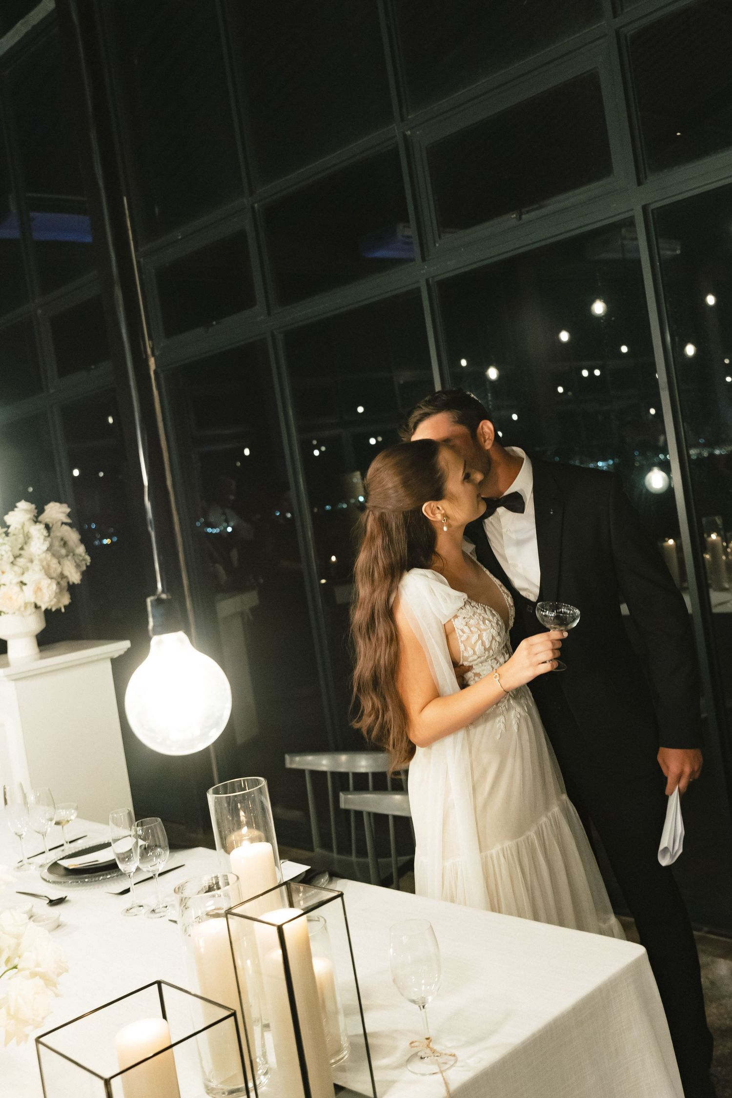 A couple in white attire shares a romantic moment at an elegant evening reception with glowing lights and white decor.