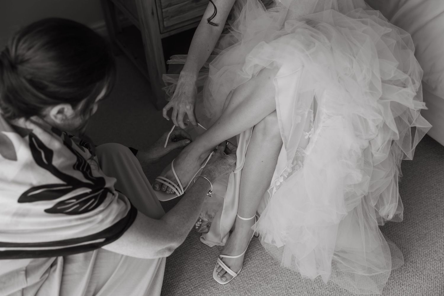 Bridesmaid helping bride put on strappy heels before wedding ceremony, black and white photo.