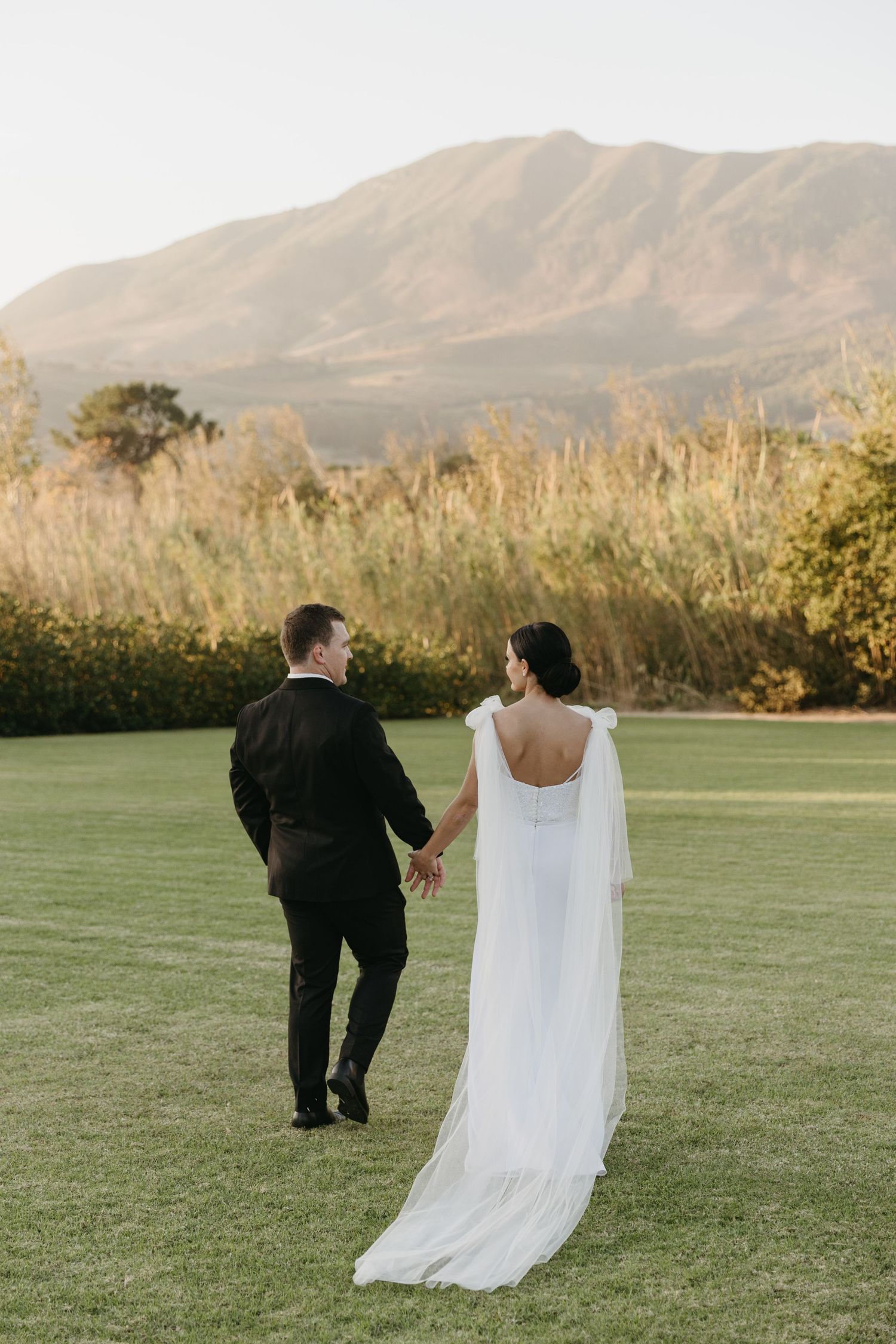 Bride and groom walking hand in hand across green lawn with mountain backdrop at golden hour wedding.